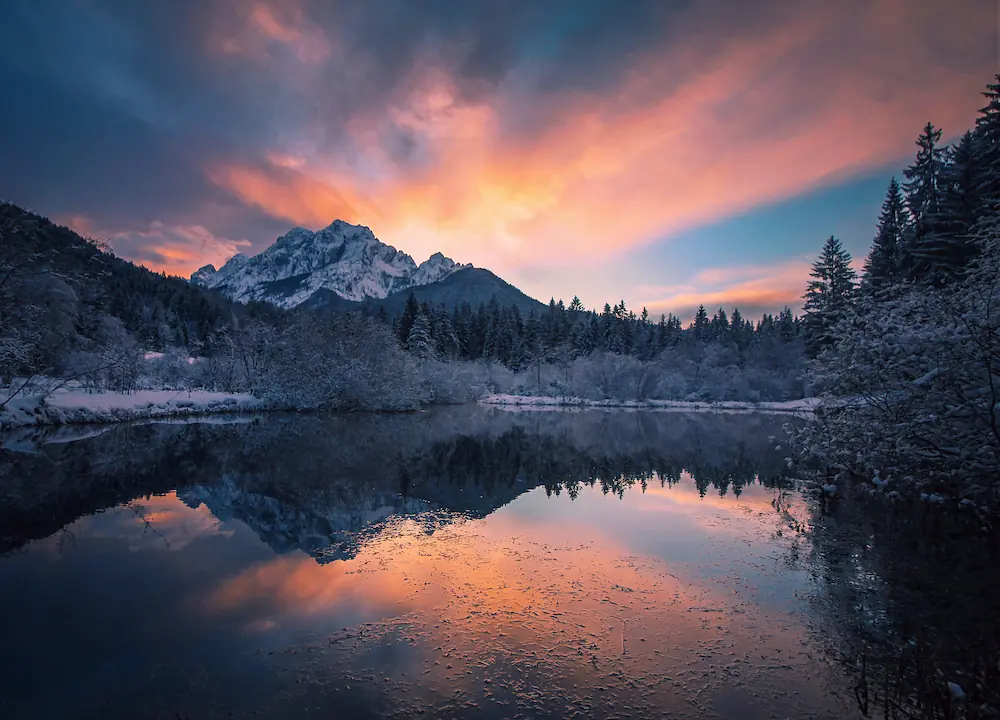 Colorful sunset reflecting in the Zelenci Springs in Slovenia, surrounded by snow covered forest and mountains.