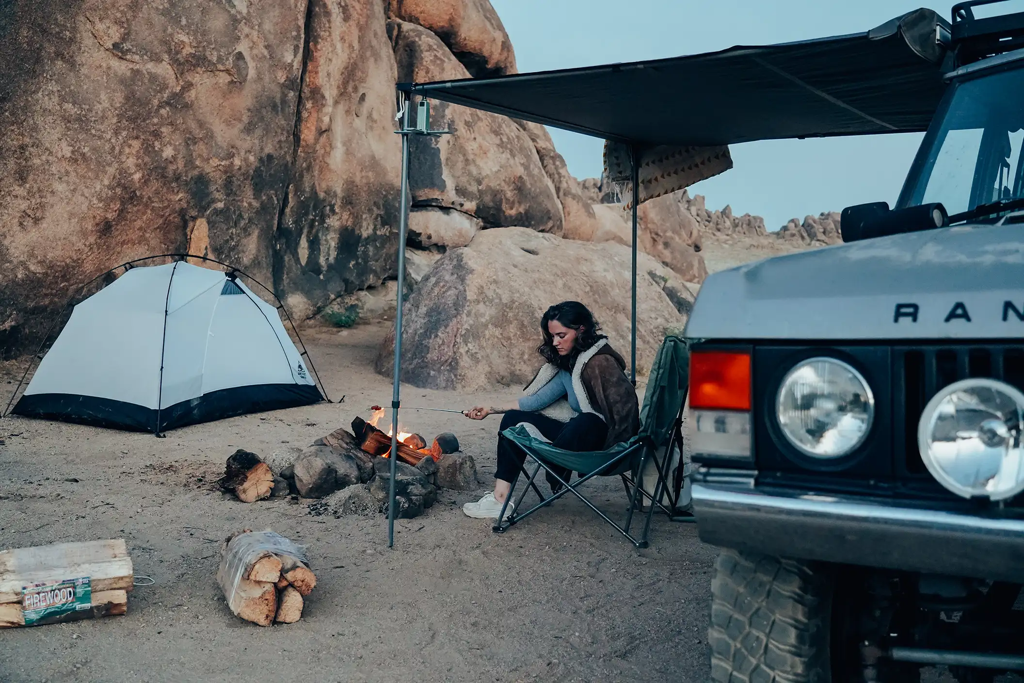 A woman cooks over a campfire, with a tent in the background and a silver Range Rover SUV in the foreground.