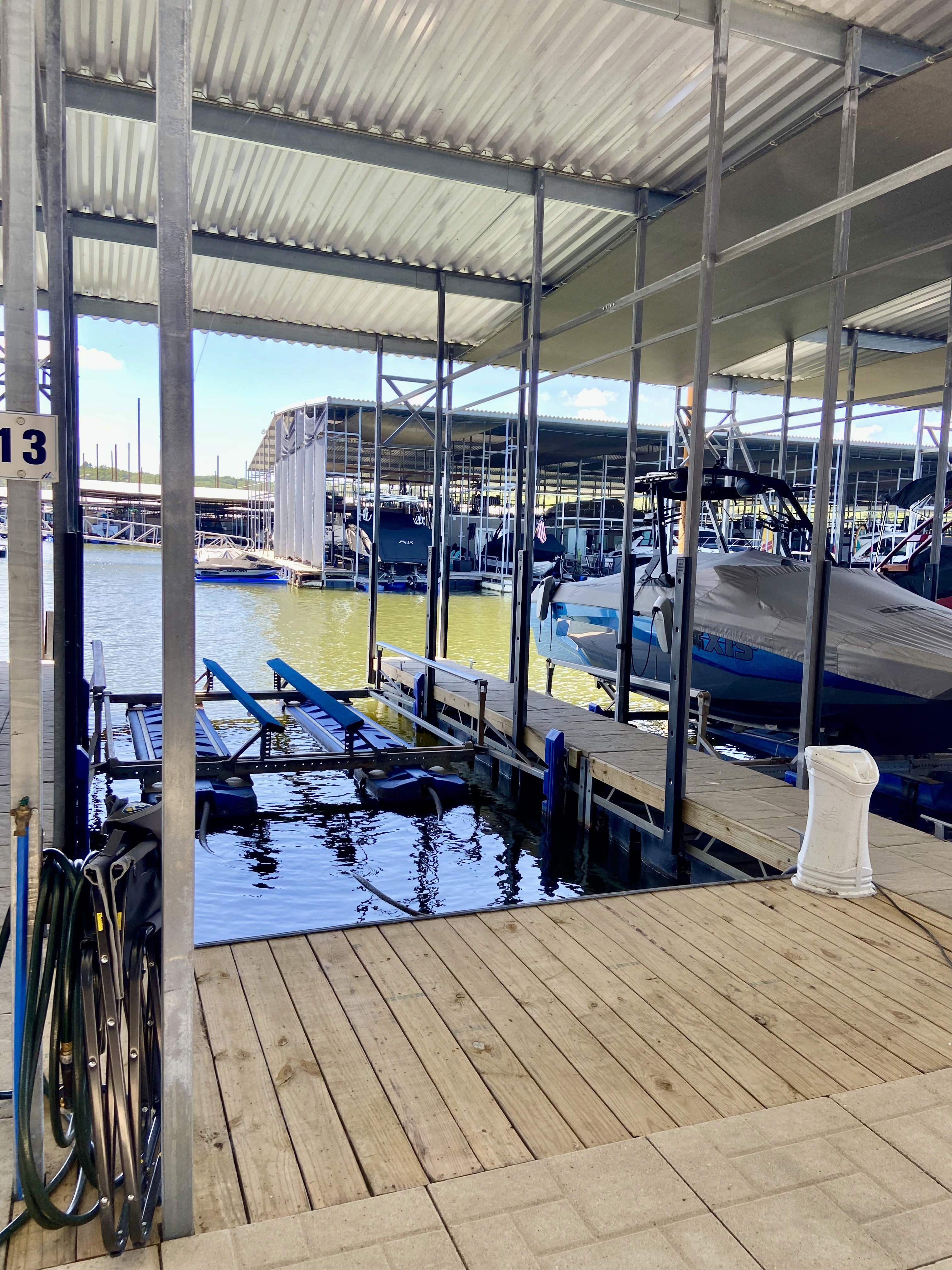 A sunlit marina with a wooden dock features several boats moored under a covered structure, with calm water reflecting the blue sky and shaded areas, creating a serene waterfront scene.