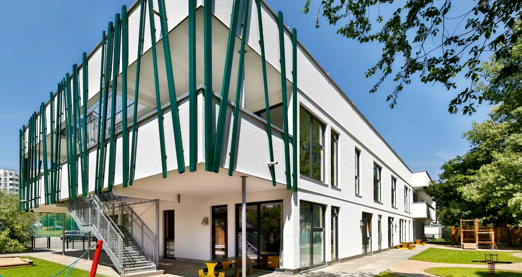 Modernes, weißes Gebäude mit grüner Fassadenverkleidung und Außentreppe im Sonnenlicht, umgeben von grüner Wiese und Bäumen.