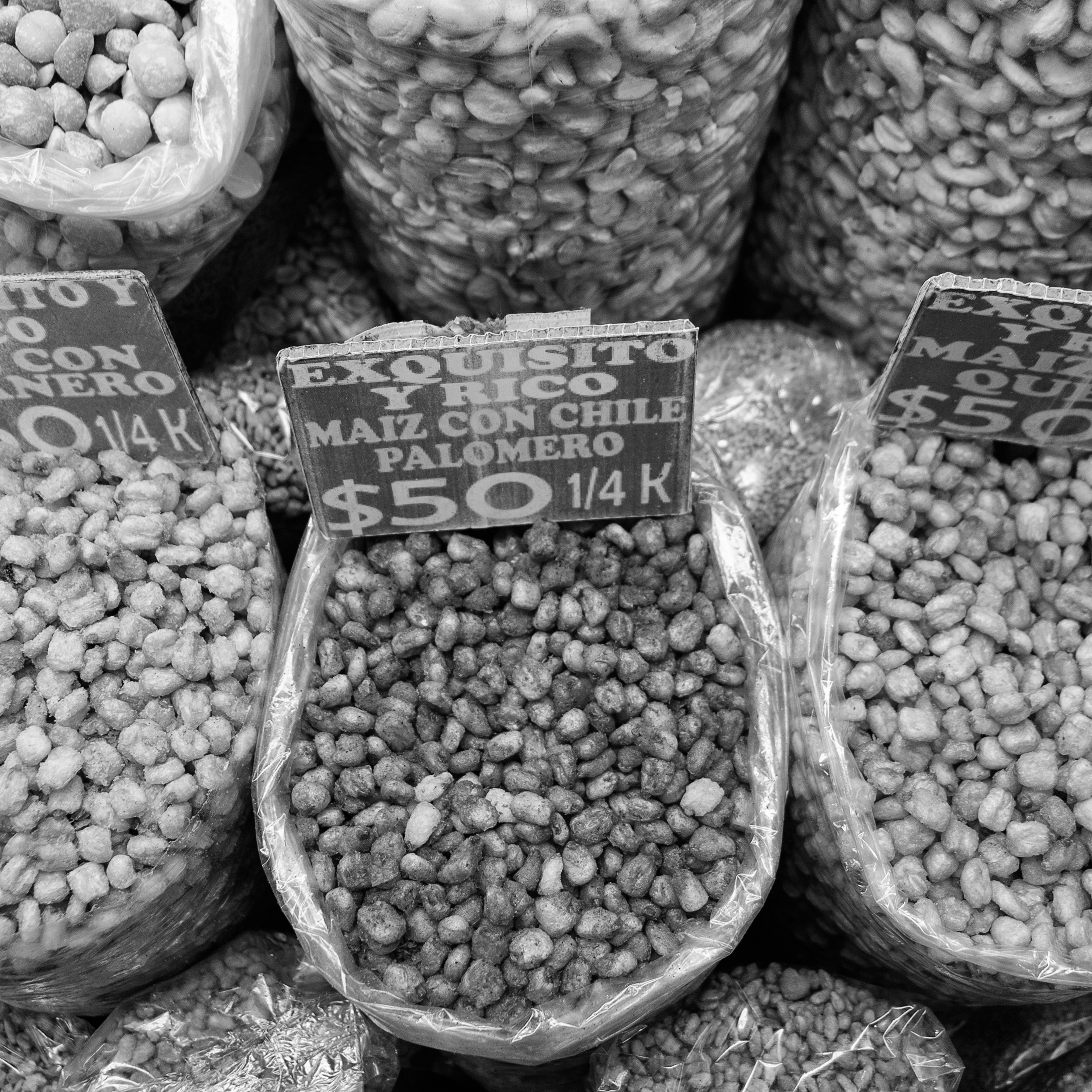 Black and white photo of market bags filled with chile-covered palomero corn, labeled with handwritten signs showing prices per quarter kilo.