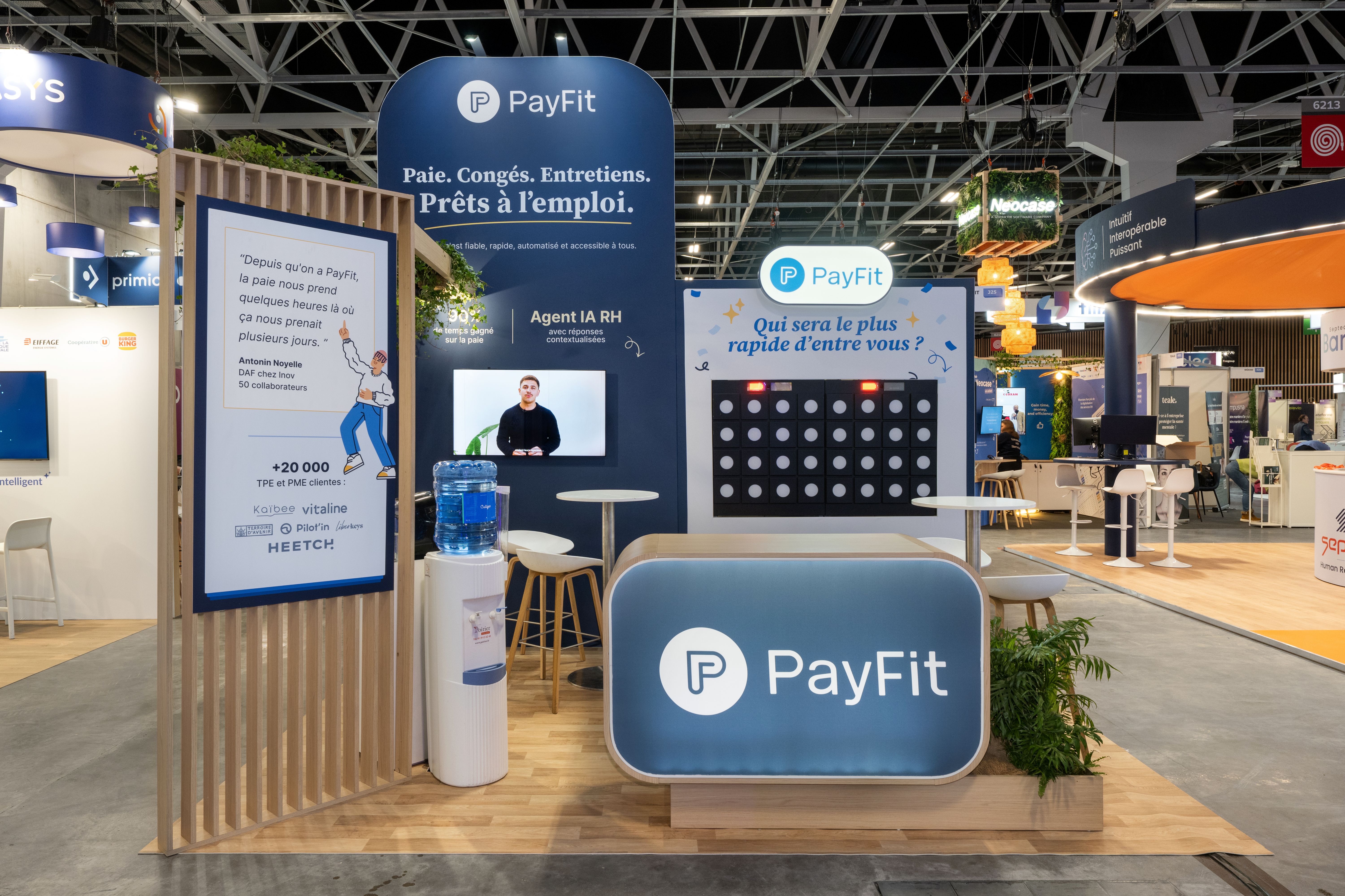 Stand PayFit salon HR Technologies 2026 réalisé par Expace – vue frontale avec arche bois végétalisée, fond bleu marine Agent IA RH, jeu interactif Puissance 4 et comptoir d'accueil arrondi bois et bleu