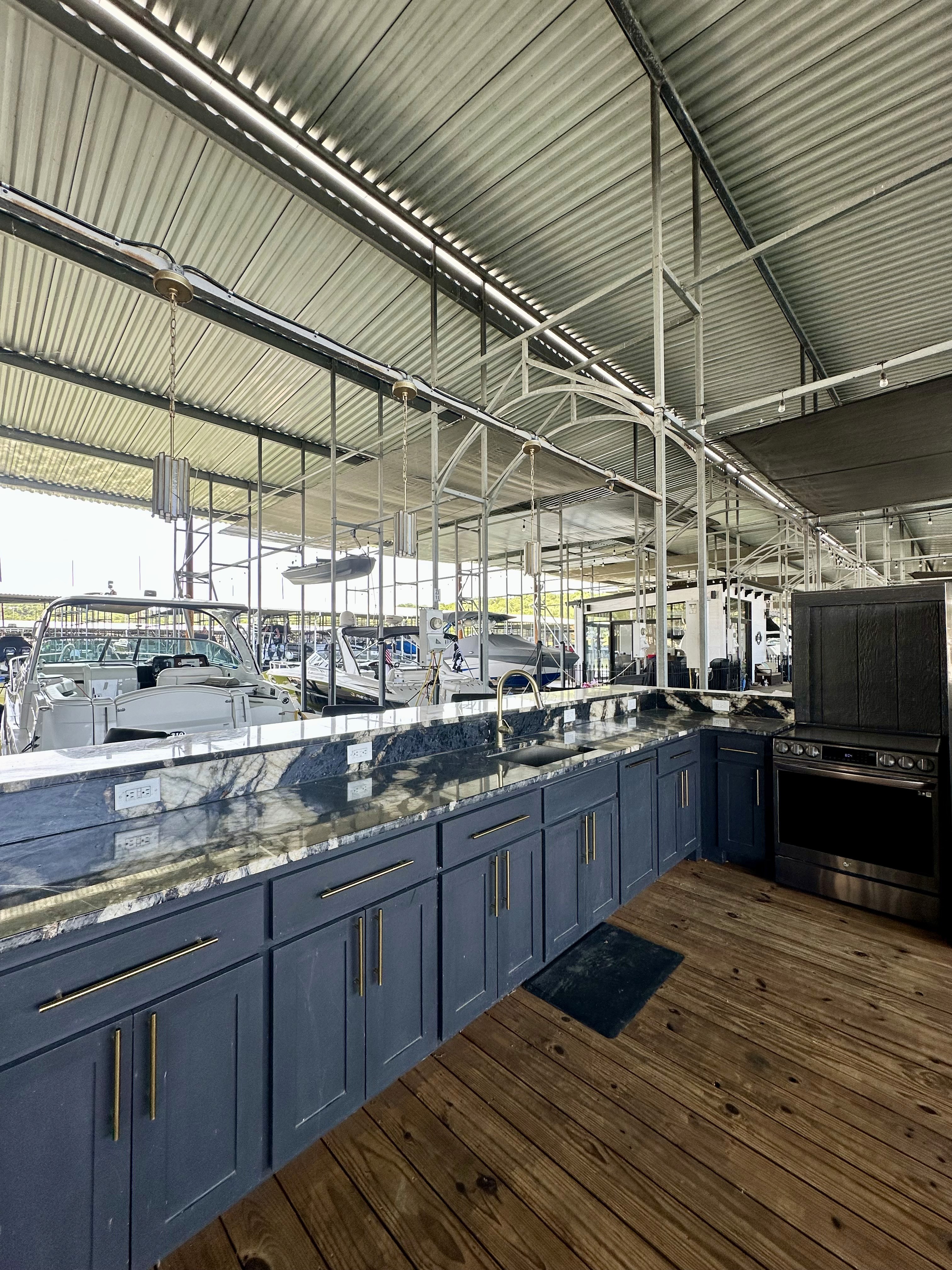 A spacious indoor boat dock featuring a row of sleek blue cabinets with marble countertops, a metallic roof structure, and several boats docked alongside a wooden floor, creating a modern and functional marina environment.
