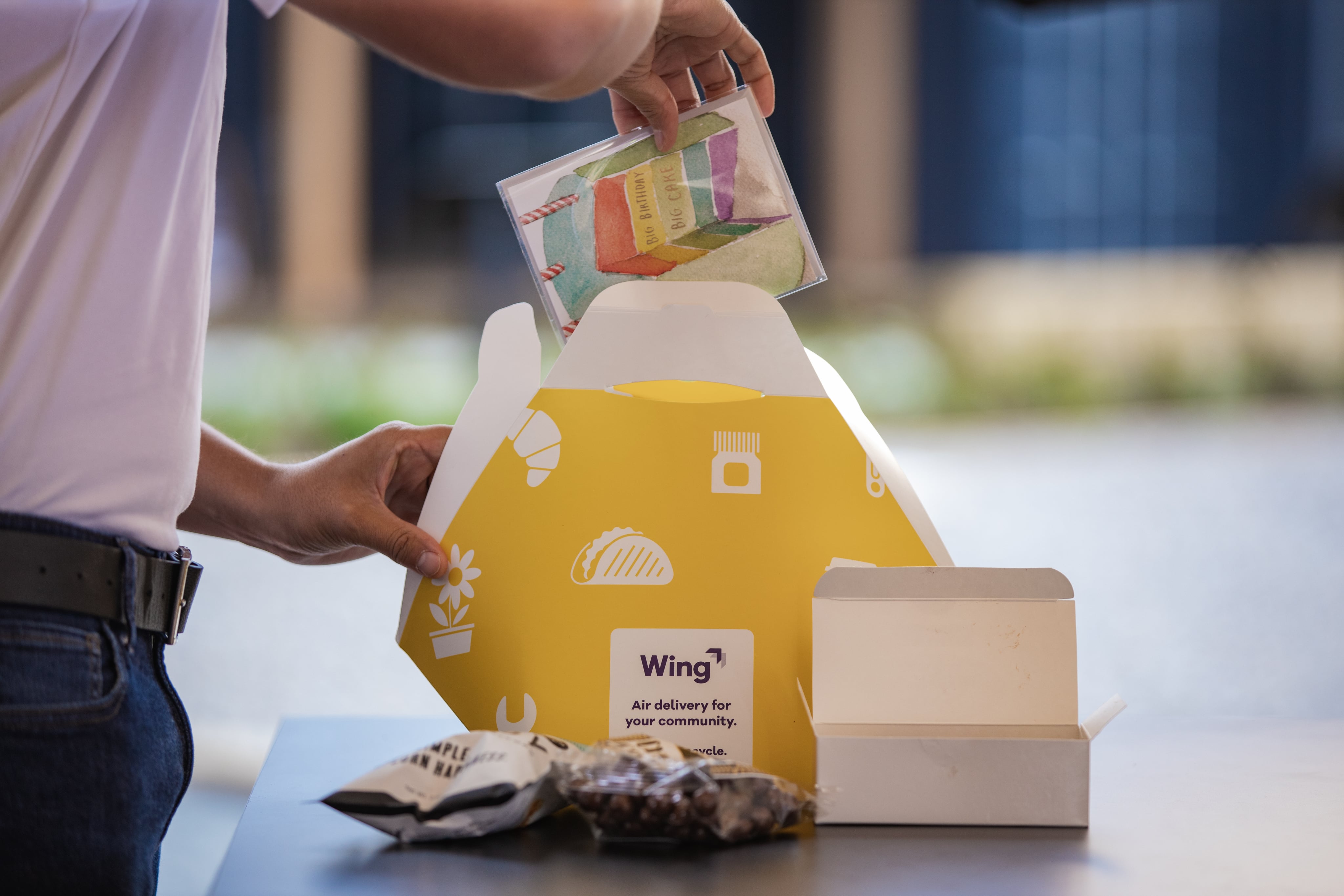 A person of-screen pulls various items out of a bright yellow Wing delivery box, including snacks and a birthday card.