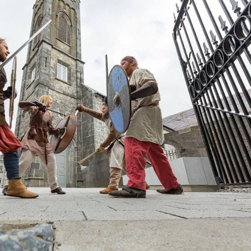 四位穿着中世纪服装的人手持剑和盾，在一座带有塔楼的石建筑外进行模拟战斗。