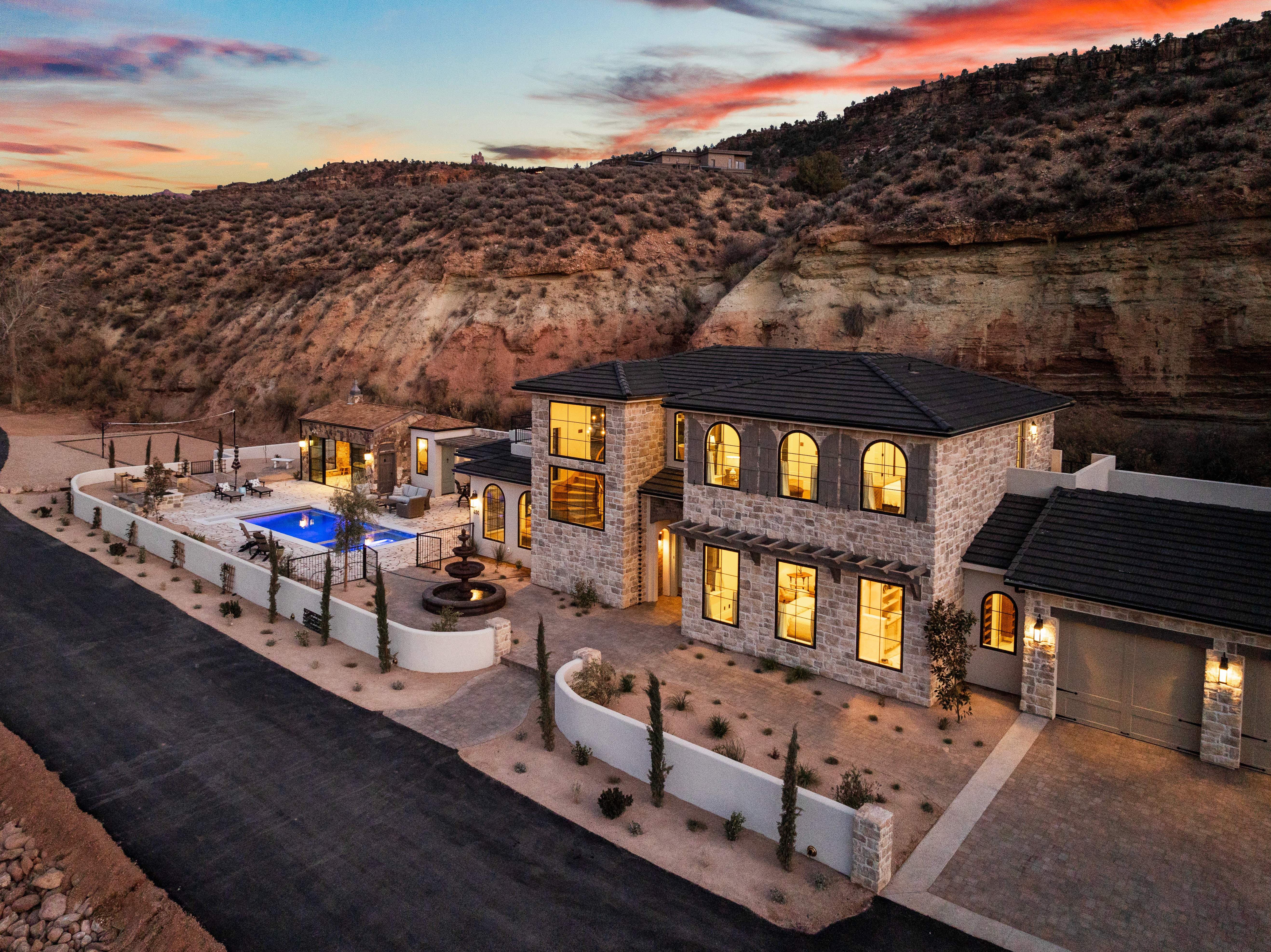 Aerial exterior view of The Cottage at Mindira Vineyard, a custom home in Rockville, Utah, featuring natural stone, arched windows, a pool, and canyon cliffs at sunset.