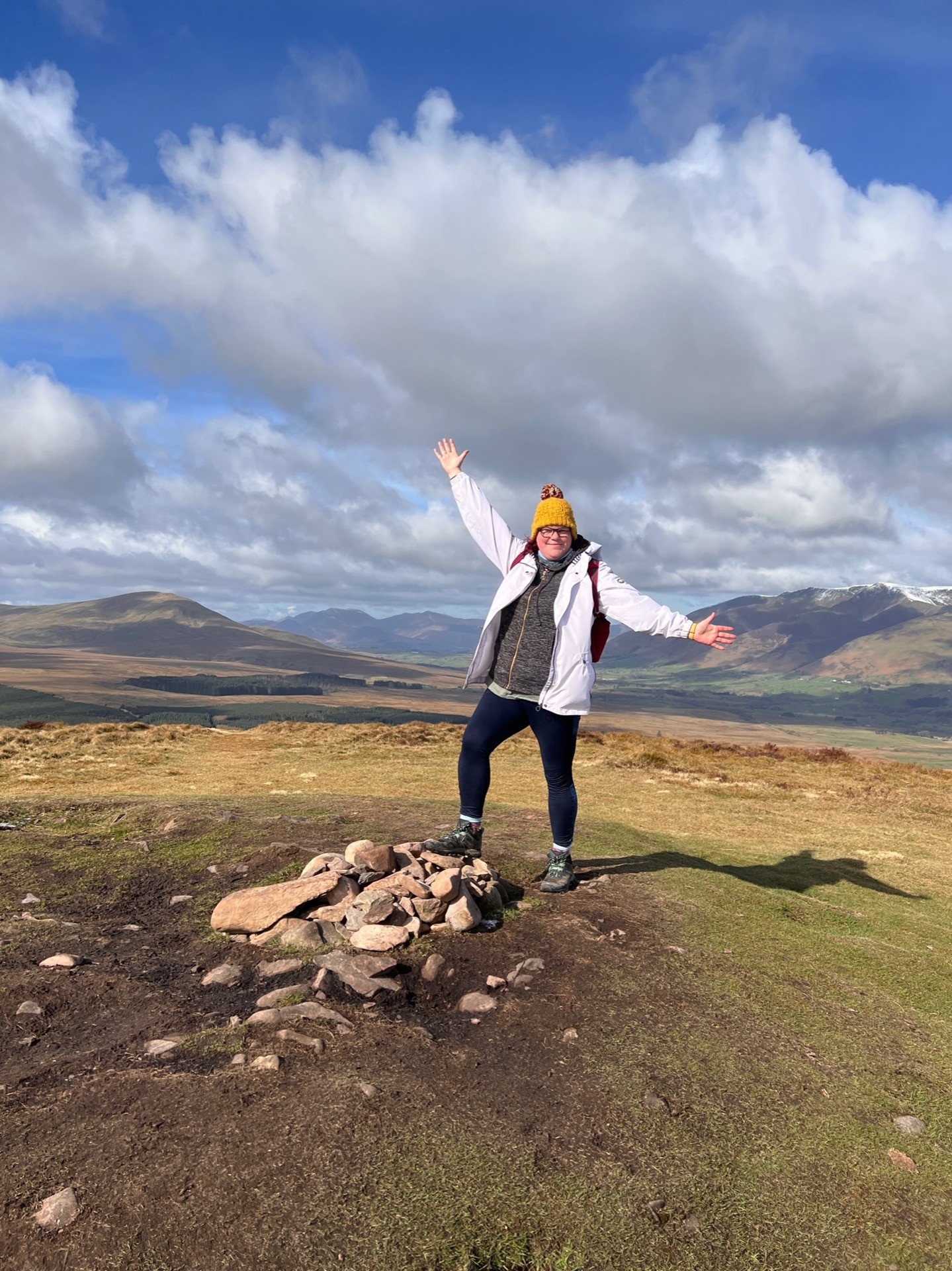 April stands with one foot on the small stone pile at the top of Great Mell Fell with her arms in the air.