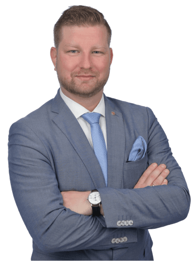 A professionally dressed man in a suit stands with arms crossed, smiling confidently against a plain background.
