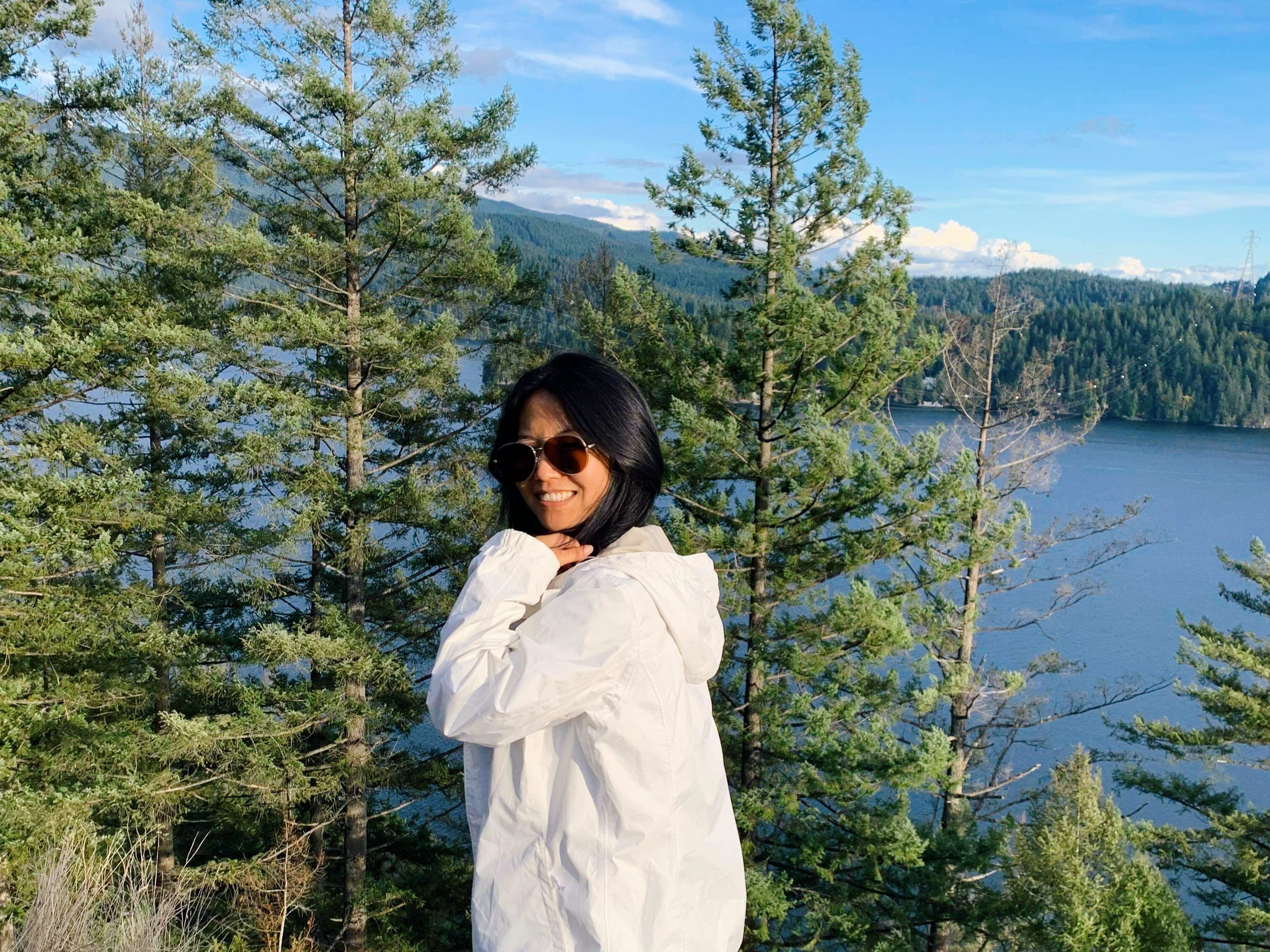 Celine: Smiling hiker enjoying a scenic forest and lake view, reflecting a love for nature and outdoor adventure.