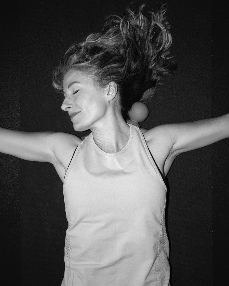 Heather Hausman lying on her back in a relaxed pose, arms outstretched, wearing a sleeveless top, against a dark background, showcasing a sense of calm and serenity.