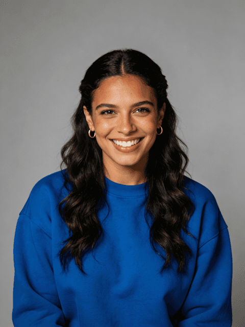 Portrait of a smiling woman with long wavy hair wearing a blue sweatshirt and gold hoop earrings against a gray background.