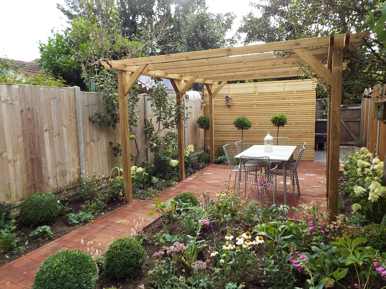 A serene garden with a wooden pergola, a table, and lush greenery along a brick path, surrounded by a fence.