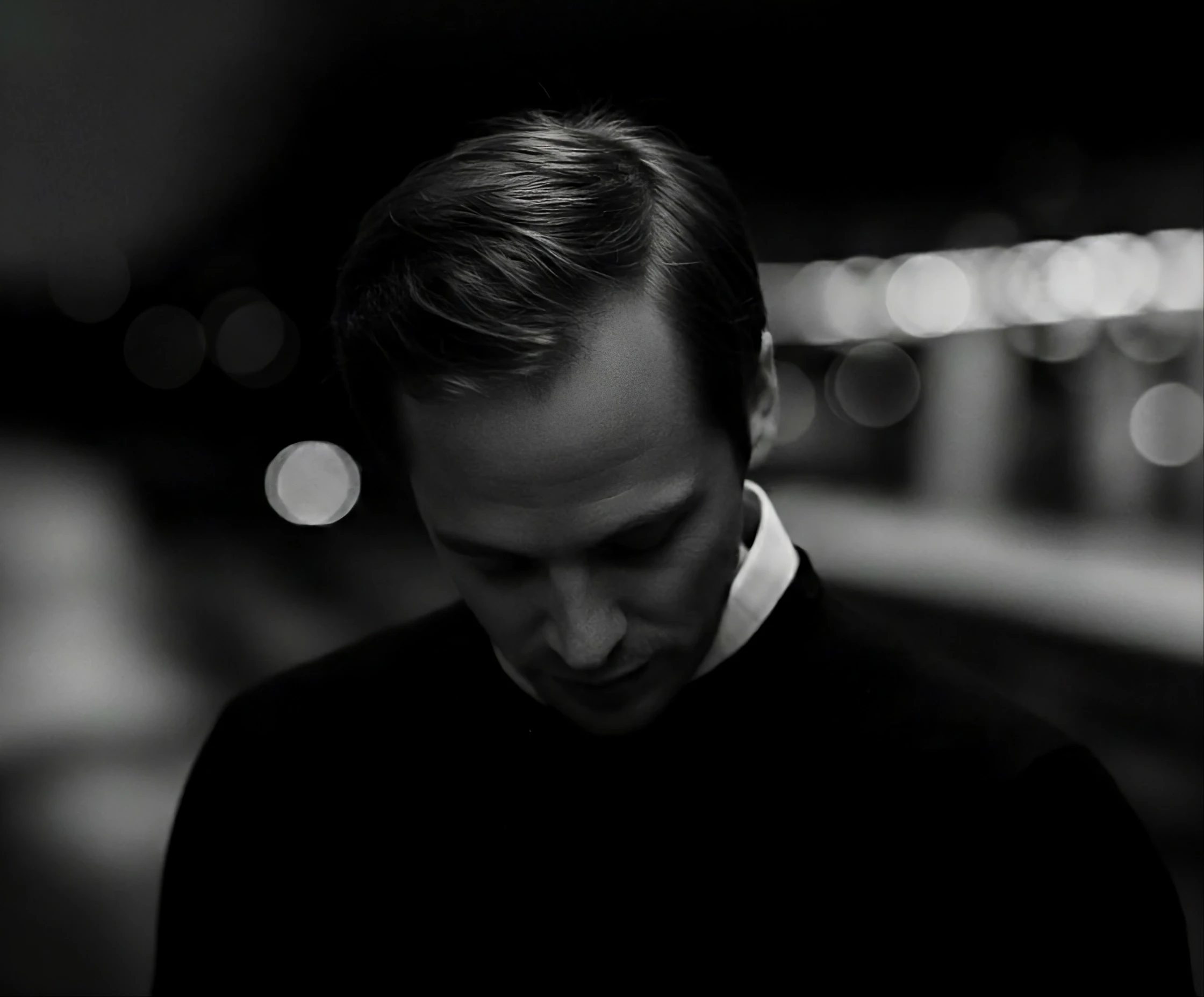 Artistic black and white portrait of a man in a dark sweater looking downward, with soft bokeh lights in the background.
