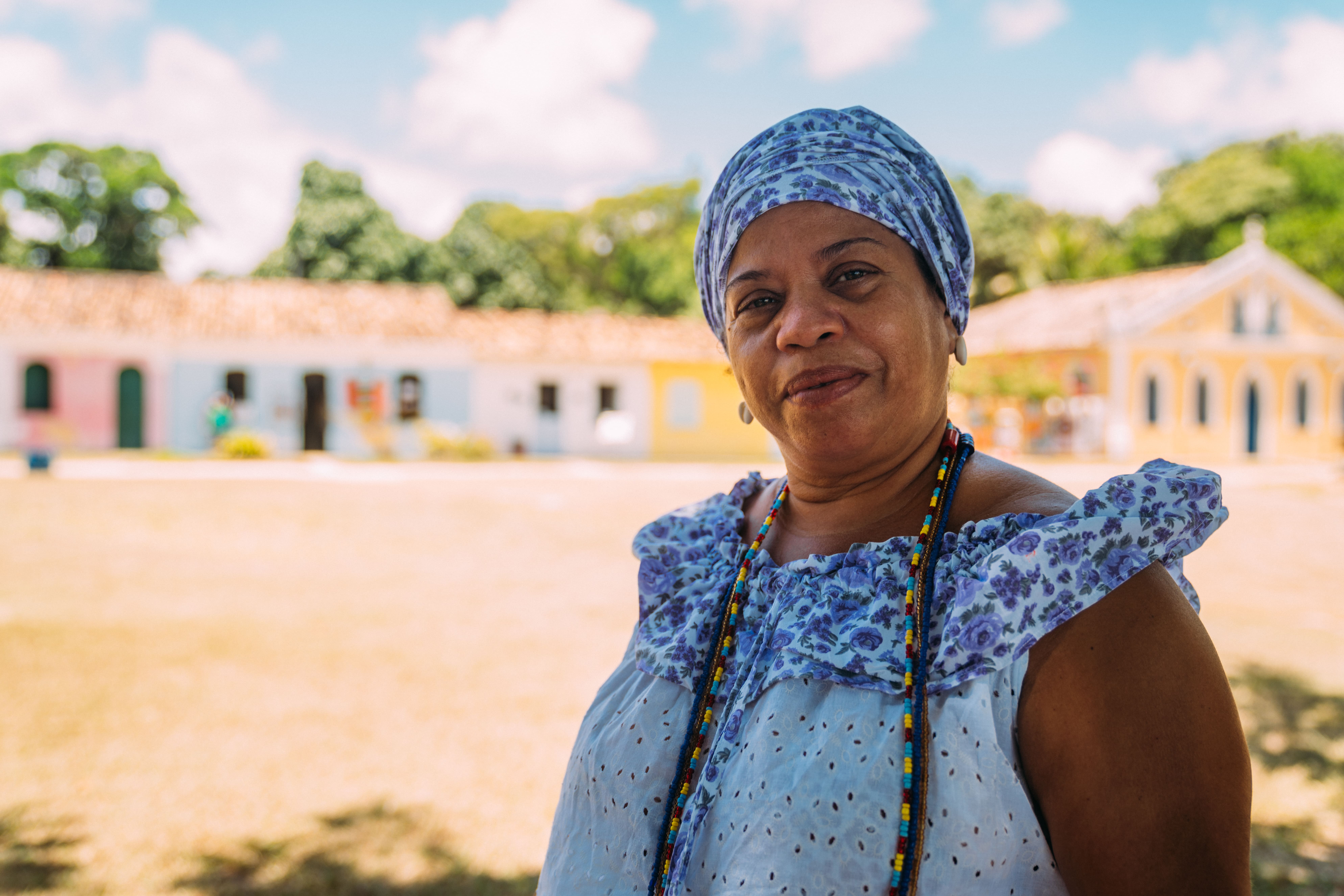 A imagem mostra uma mulher sorridente vestida com um traje tradicional baiano, característico da religião Umbanda. Ela está ao ar livre, em frente a um cenário que parece ser o centro histórico de Porto Seguro, com casas coloniais coloridas ao fundo e um céu claro. A mulher usa um turbante estampado e um vestido com detalhes florais em tons de lilás e azul, além de colares de contas coloridas, típicos das tradições religiosas afro-brasileiras. A luz natural ilumina a cena, trazendo uma sensação de tranquilidade e orgulho cultural.