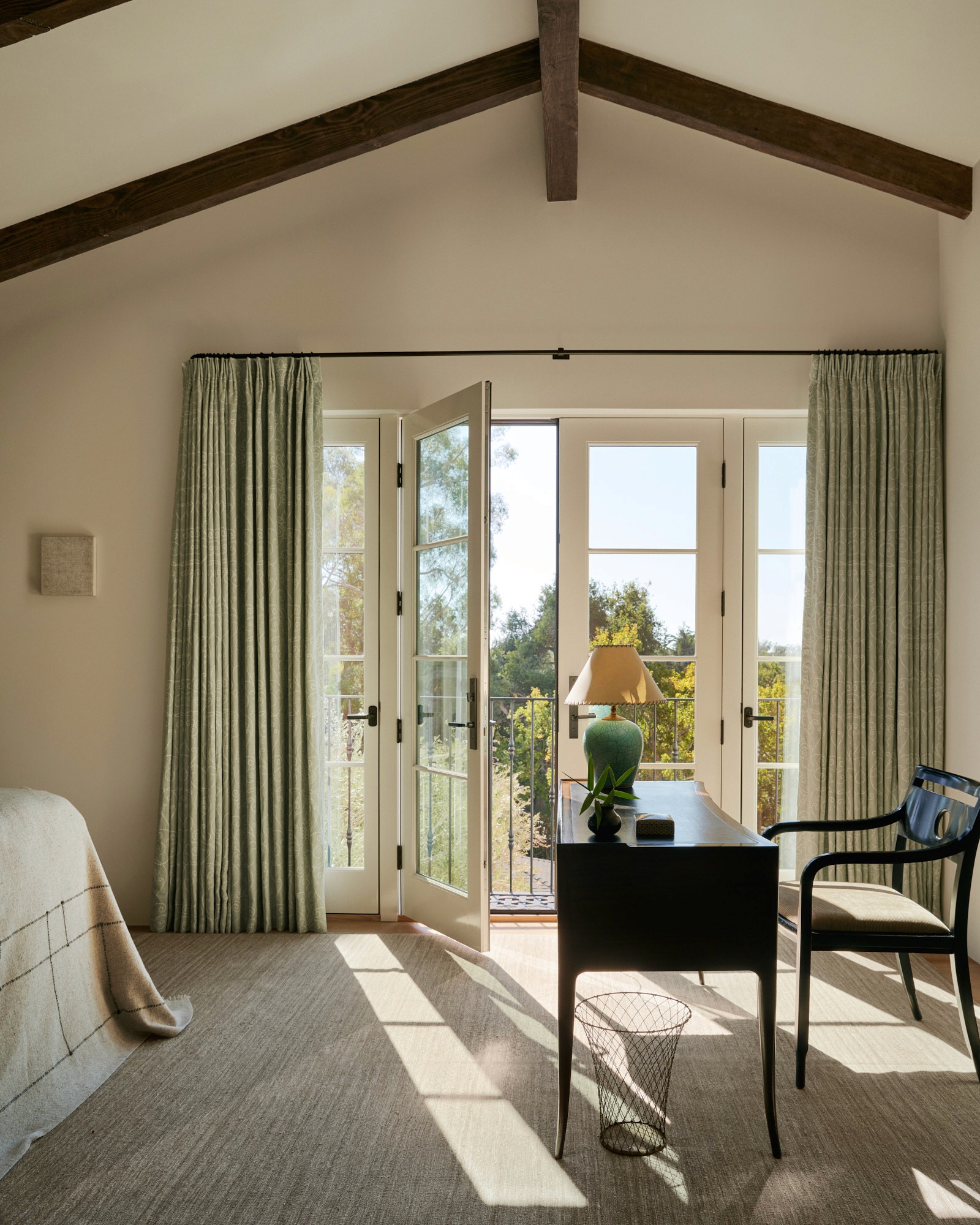 A vaulted bedroom with exposed ridge beam and French doors open to a balcony. A dark writing desk and blue-black chair are positioned to catch the light. Sage green drapery frames the doors, and a small plaster sconce is mounted on the far wall.