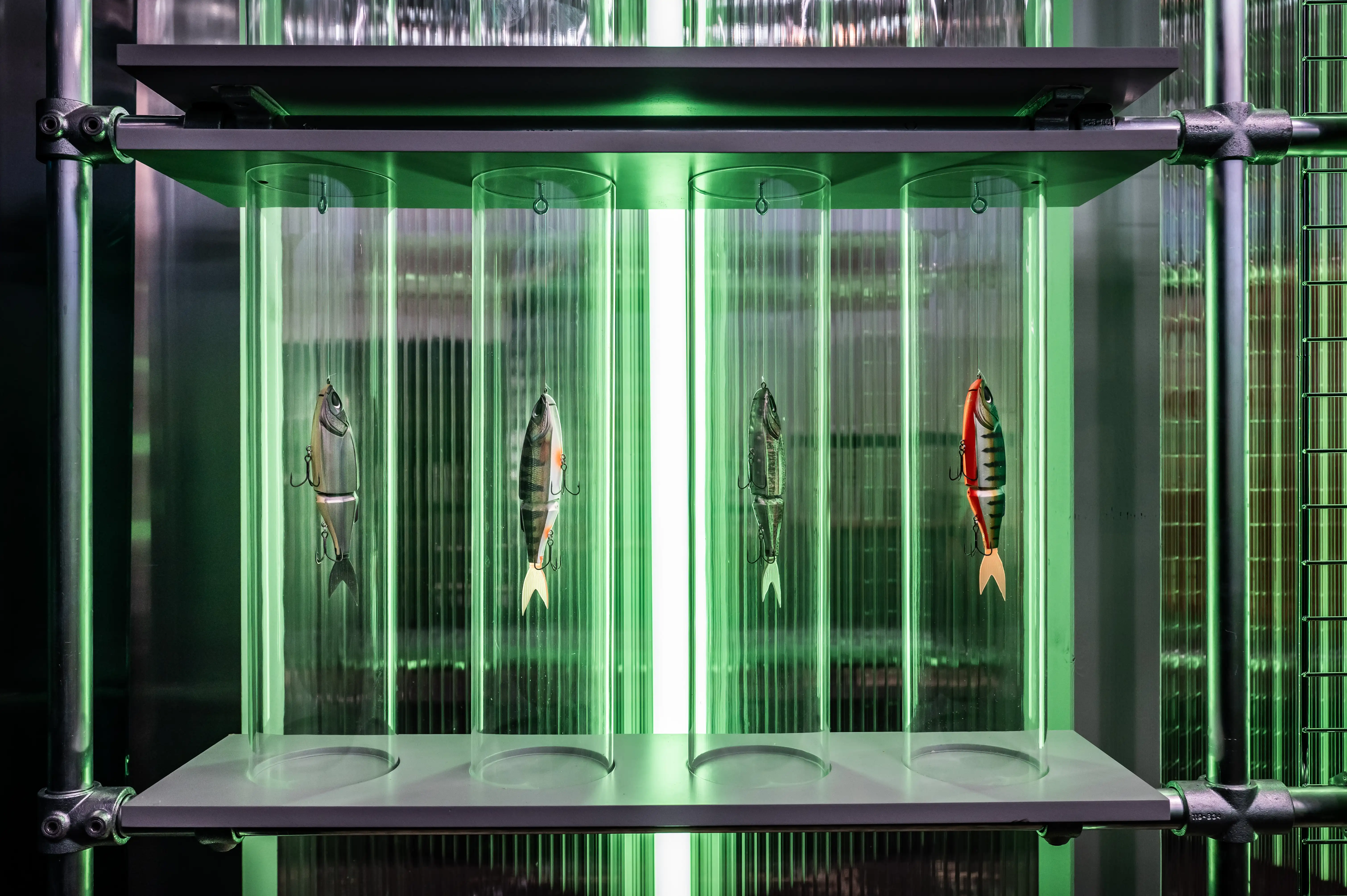 Long metal facade of the Nays booth with glowing logo and green-lit glass cylinders on a pebble bed.