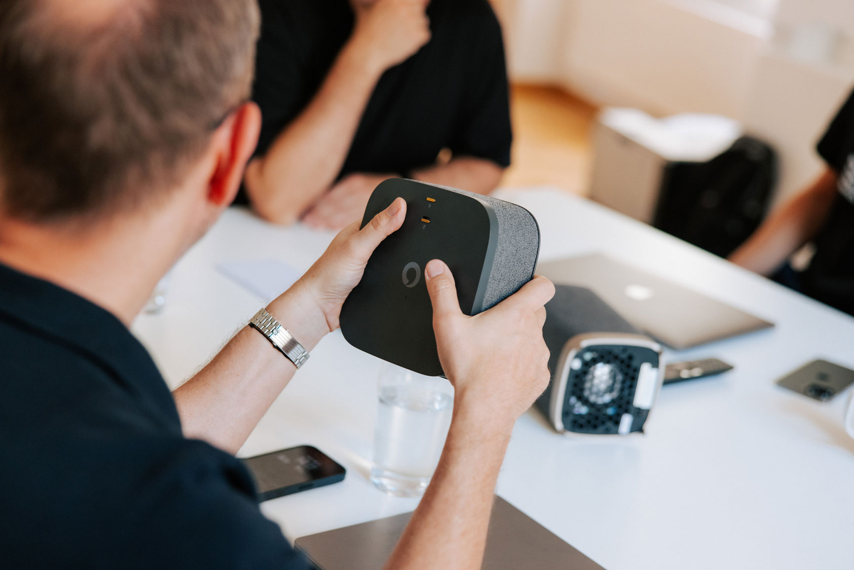 A person holds a prototype of a TV set-top box during a workshop, while colleagues sit around a table reviewing industrial design concepts in a bright office setting