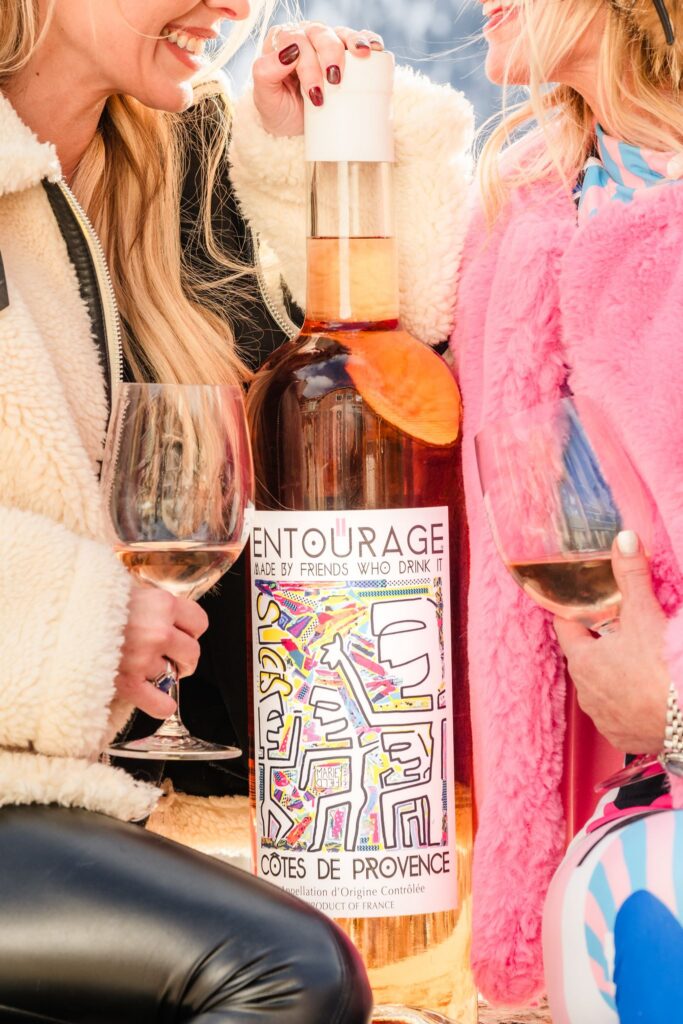 Two guests in winter coats holding wine glasses beside an oversized Entourage rosé bottle