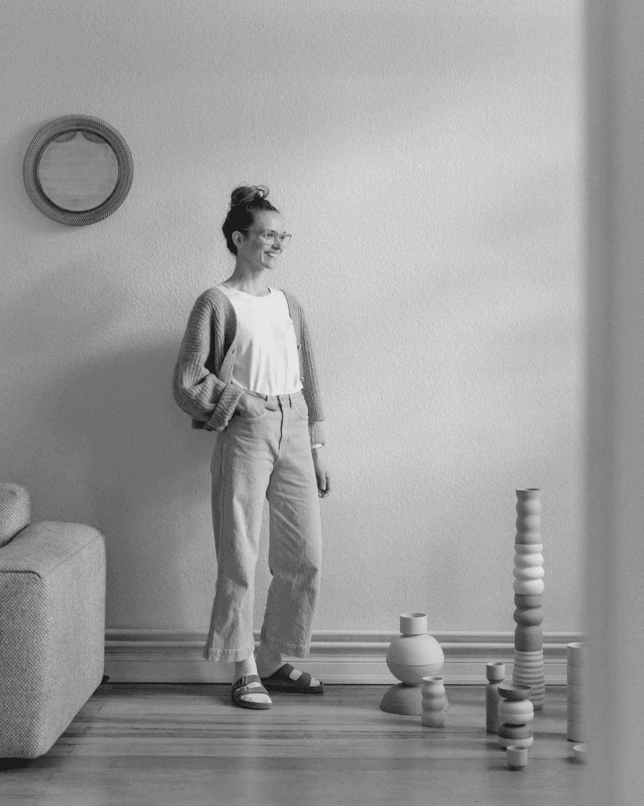 A person stands in a minimalist living room with a circular wall mirror, wearing casual clothes, next to a stack of artistically arranged, geometric vases and pillars on a wooden floor.