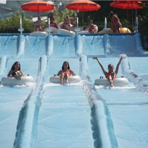 Three people on inner tubes slide down parallel water slides, with others waiting at the top under red umbrellas.