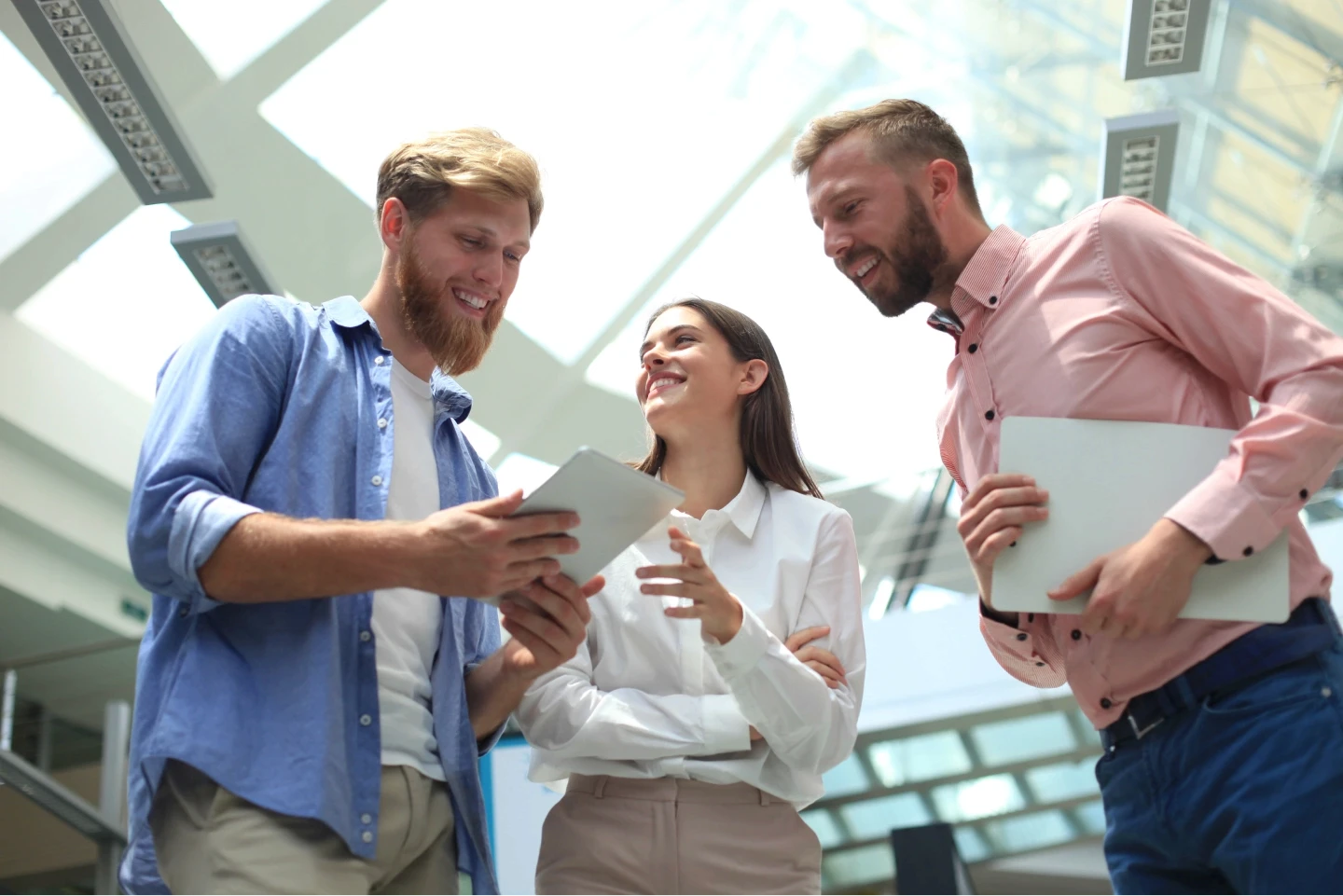 Three people are engaged in a discussion, smiling and looking at a tablet inside a bright, modern office space with large windows and contemporary architecture.