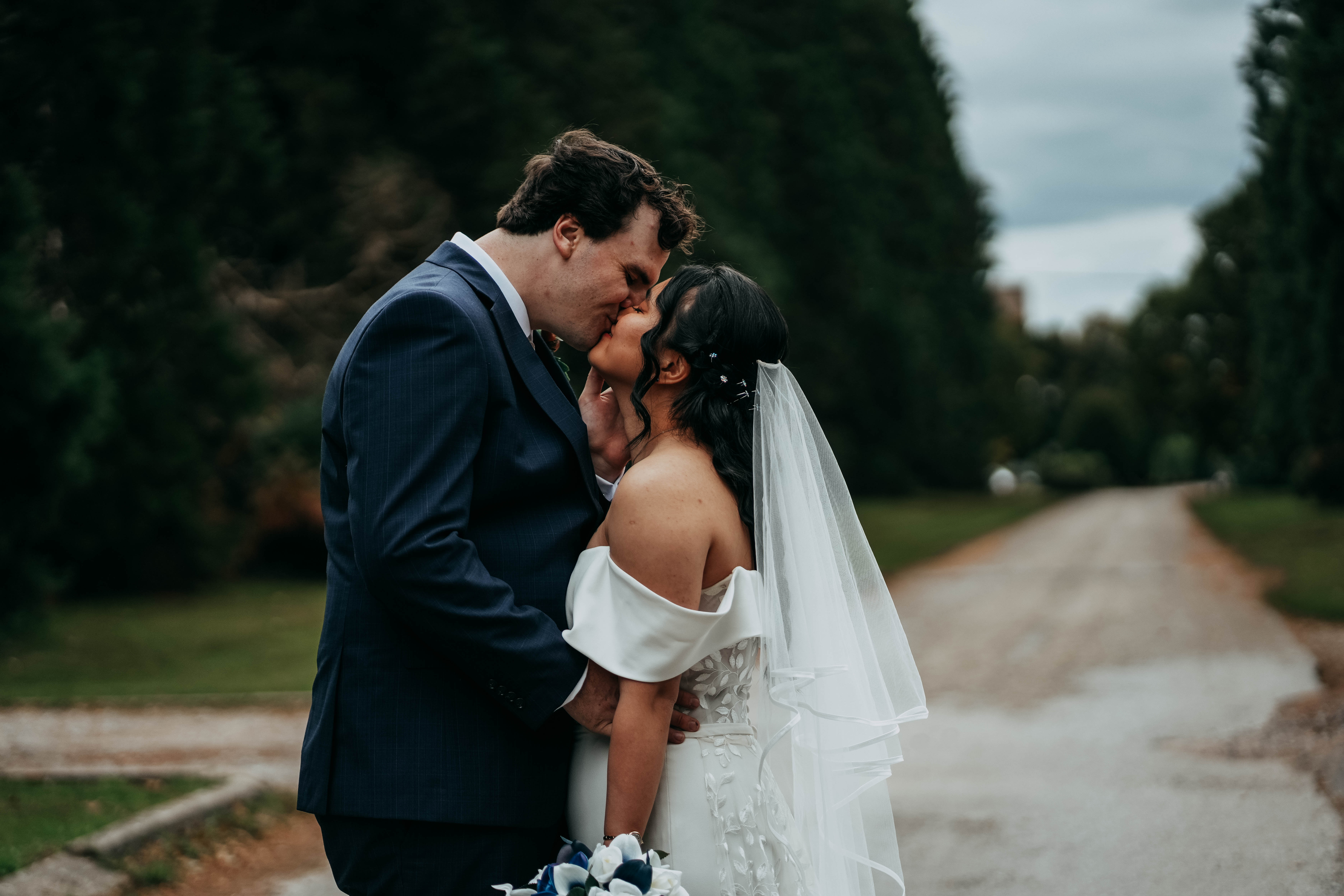 A close-up, romantic photo of the bride and groom sharing a kiss on a tree-lined road. The groom gently cups the bride's cheek with his hand as they embrace.