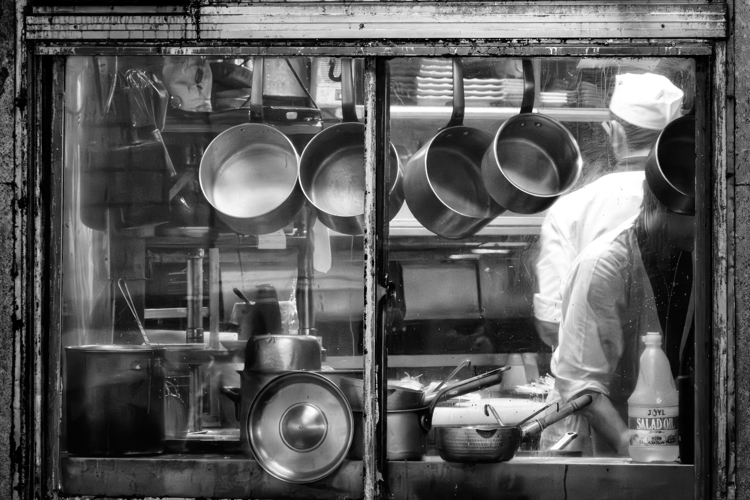 Black and white photograph of kitchen pots and pans hanging in front of a window while two cooks work inside.
