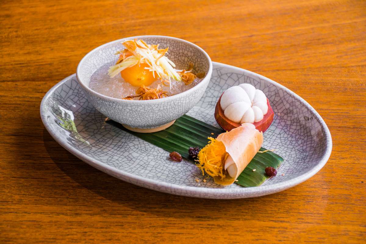 A bowl of orange and white soup with shredded toppings sits on a large plate next to a small dumpling and garnish.