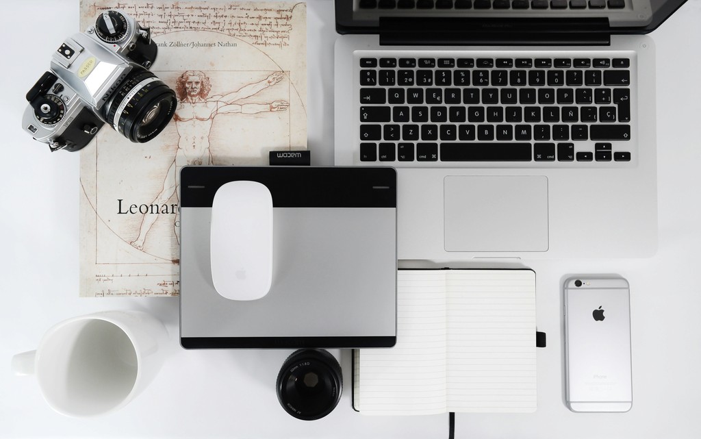 A modern workspace setup featuring a MacBook, a Wacom tablet with a mouse, an iPhone, a vintage camera, an open notebook, and a coffee mug, all arranged on a white desk alongside a Leonardo da Vinci illustration.