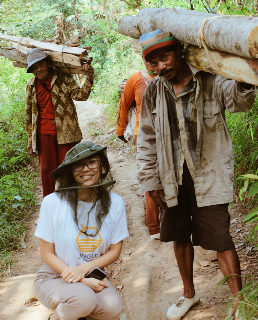 A woman in a white t-shirt and a mesh hat smiles while kneeling on a dirt path. Two men are seen behind her, with one carrying a bundle of firewood on his back and the other standing next to her. The scene is set in a lush, green environment.