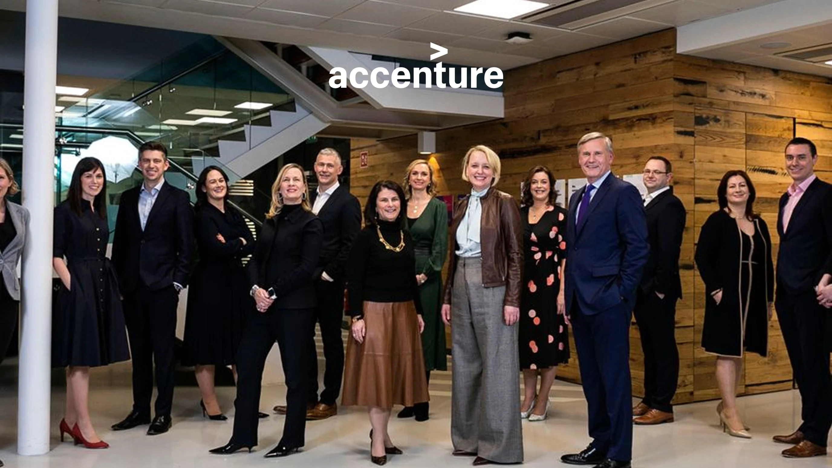 Accenture team photo featuring a large group of professionally dressed consultants posed indoors in a modern office with the Accenture logo overhead