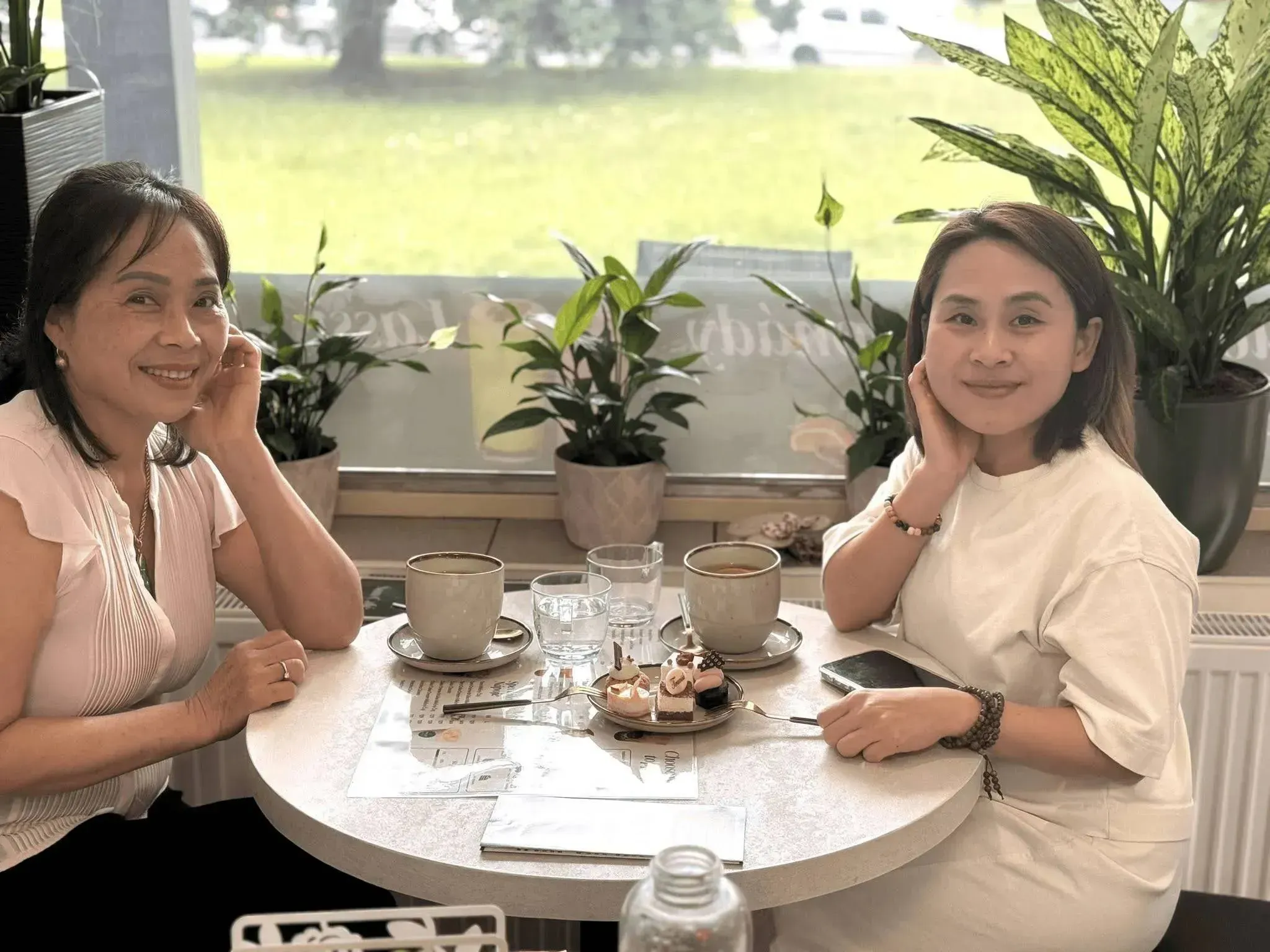 Two women smiling and enjoying coffee and a small cake at a bright café table surrounded by plants