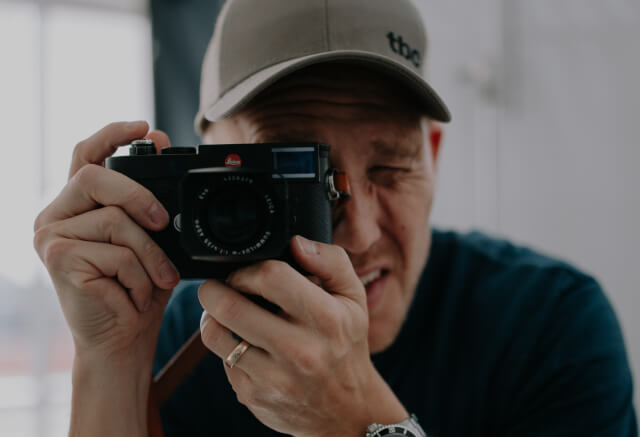 A person wearing a cap holds a camera up to their face, looking through the viewfinder as if taking a picture.