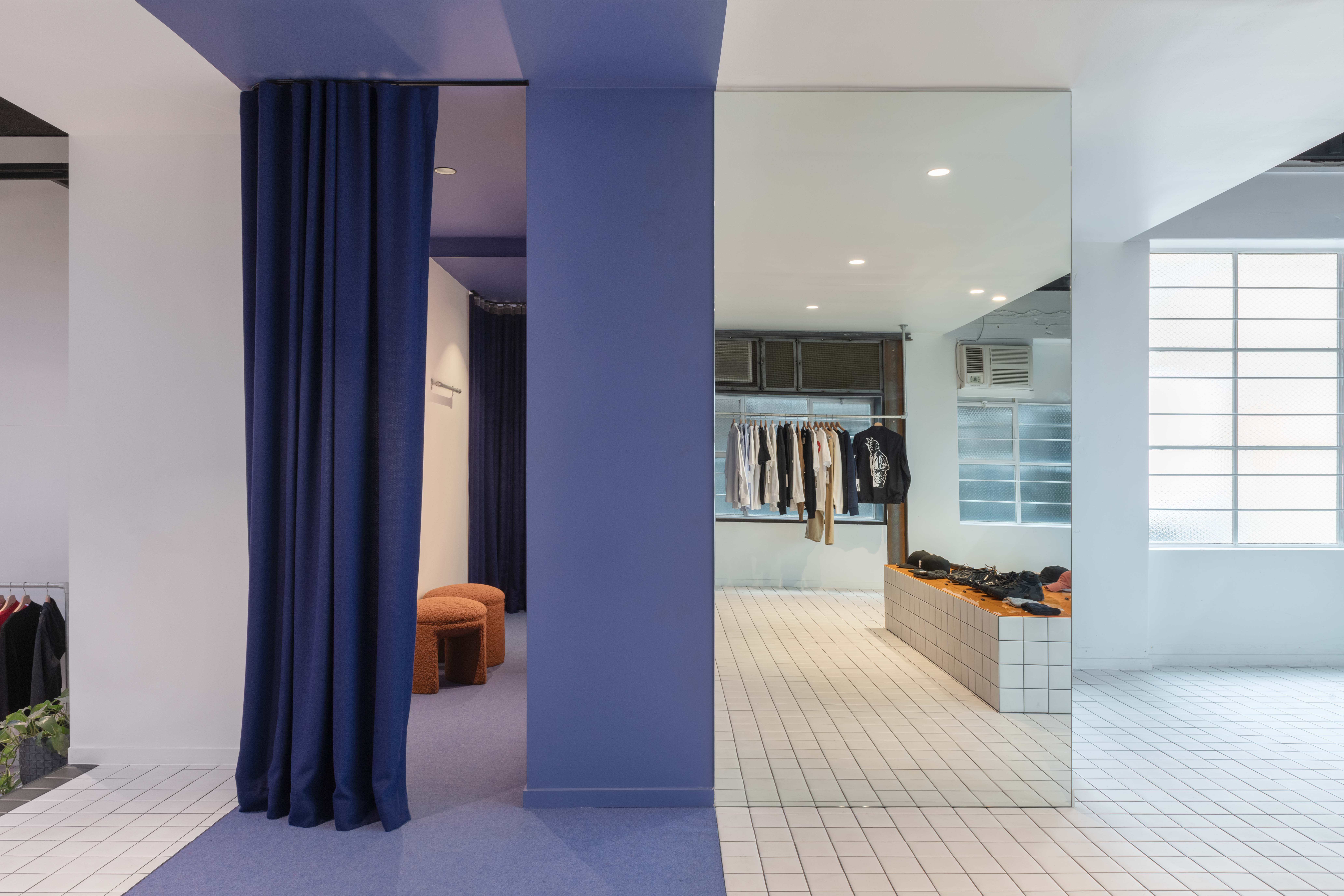 Split interior of the 199A® space showing contrasting zones, with deep blue walls and curtains transitioning into a bright tiled retail area with clothing rails and minimalist fixtures.