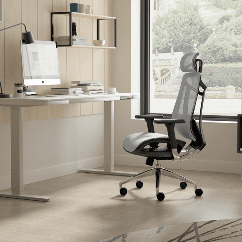 Lifestyle shot of a grey mesh ergonomic office chair in a bright, modern home office setting with a white standing desk, computer, and natural window light.