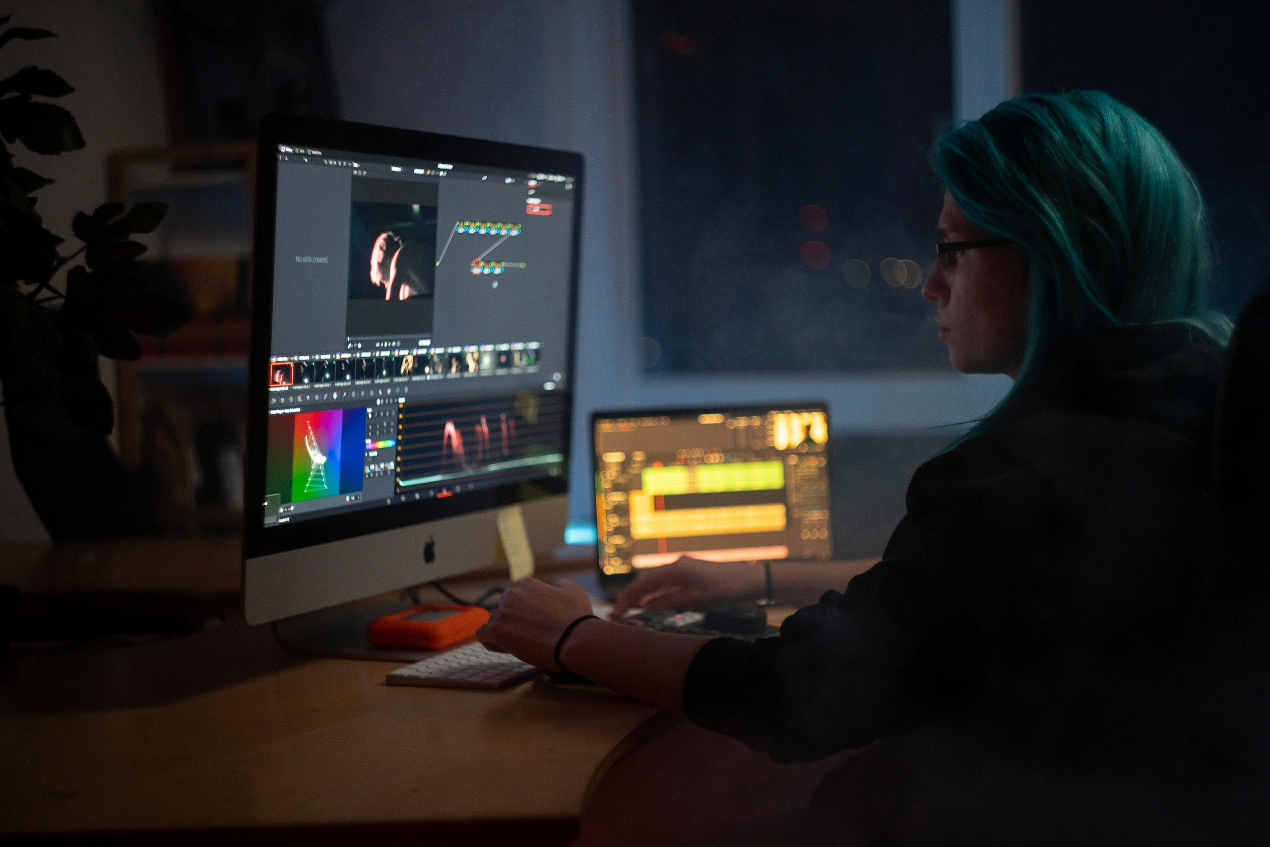 Man in a cap and glasses working at a wooden desk with documents and a laptop. The dimly lit room has a focused, thoughtful atmosphere.