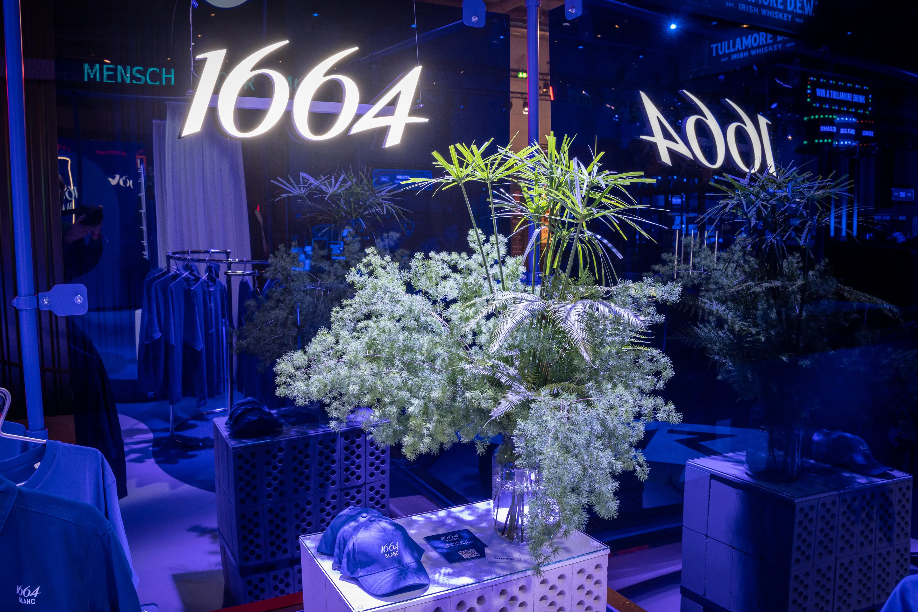 Delicate greenery in a glass vase on a white crate table in front of the glowing 1664 lettering in the blue-lit lounge.