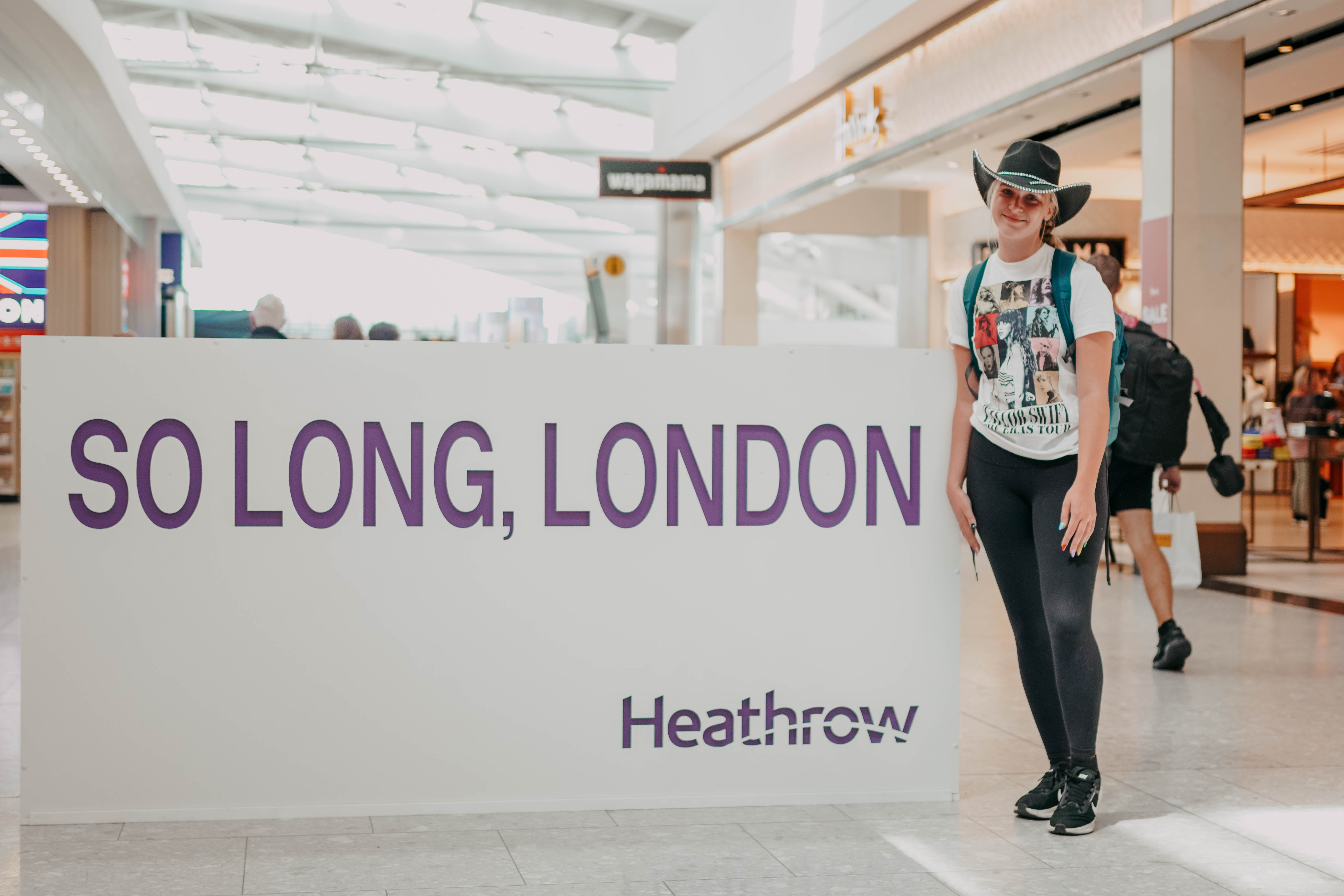 A young woman wearing a Taylor Swift tour t-shirt and a black cowboy hat smiles for a photo in a bright airport terminal. She is standing next to a large white sign with purple text that reads, "SO LONG, LONDON" and "Heathrow."