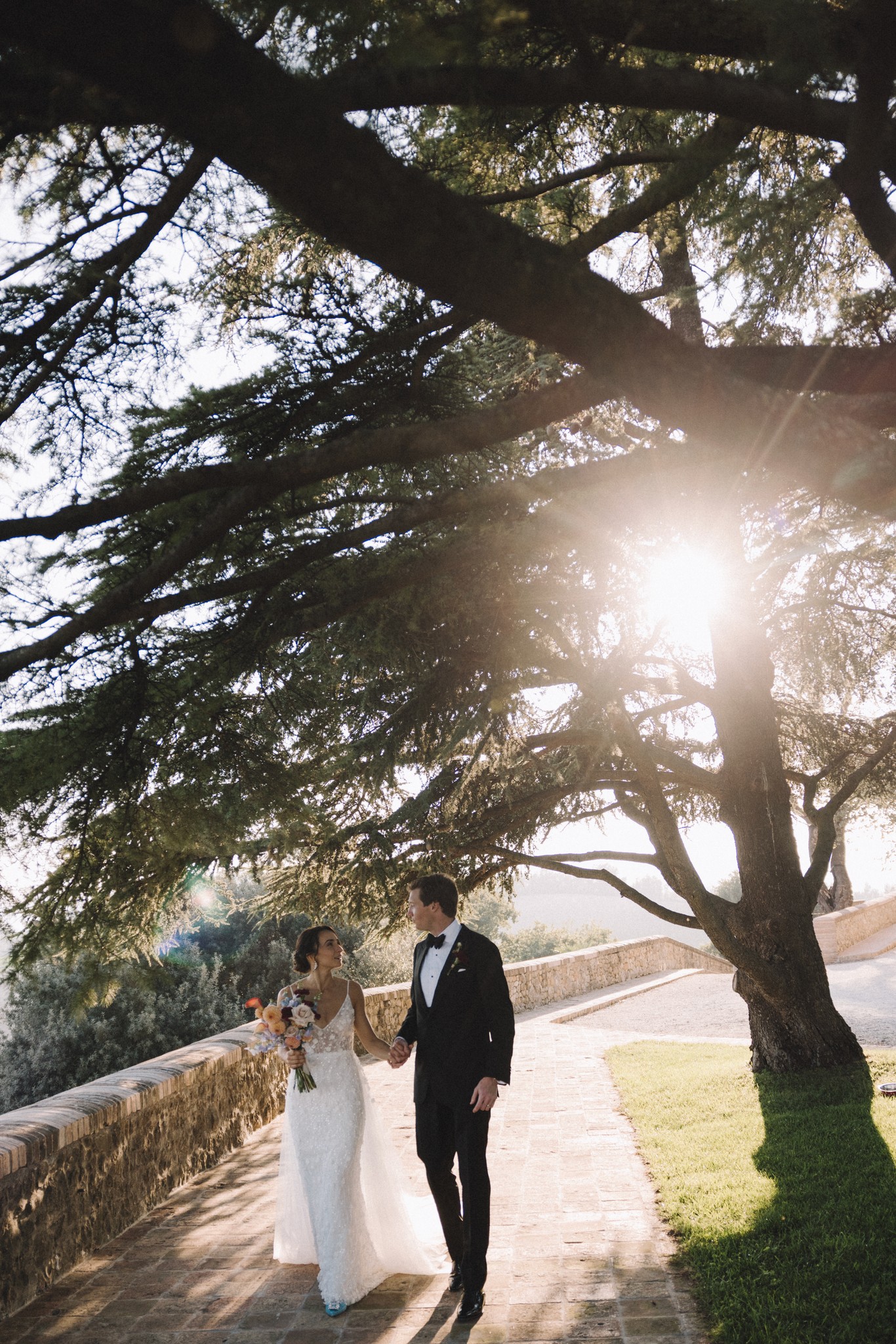 Sposi camminano sotto alberi monumentali al Castello di Tabiano, luce dorata tra i rami nel reportage matrimoniale