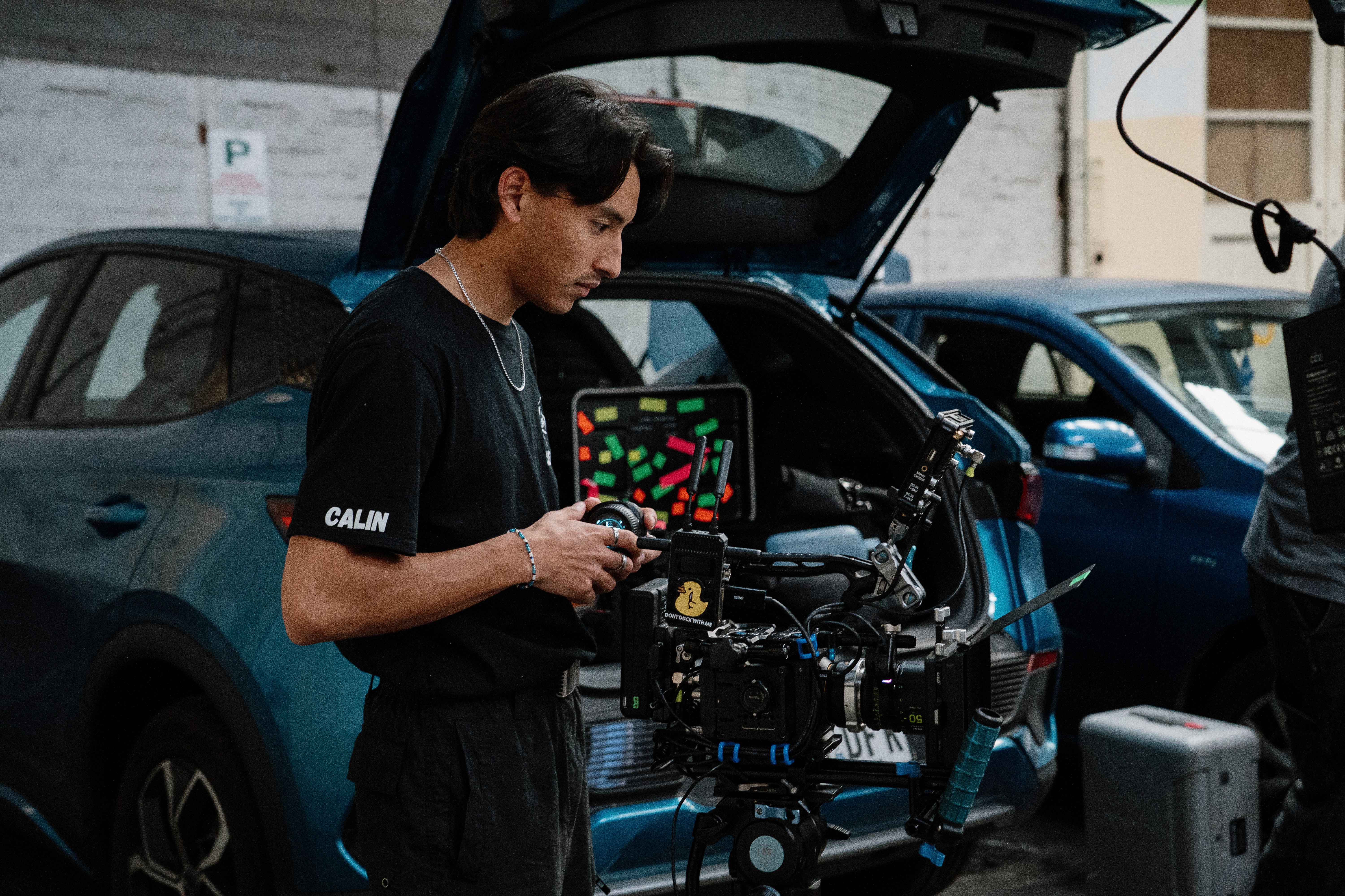 Two camera operators in a dimly lit studio adjust a large video camera. They wear headsets and caps, focusing intently. Mood is professional and focused.