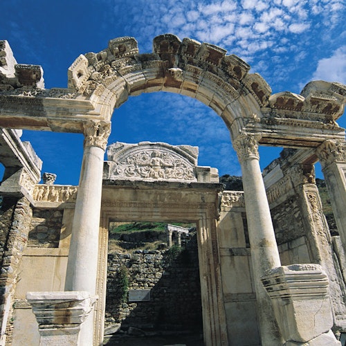 Ancient stone archway with ornate carvings, flanked by tall columns, against a partly cloudy blue sky.