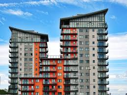 A view of three modern, multi-story residential buildings set against a bright blue sky with white wispy clouds. The two taller towers are primarily grey and white, while a connecting, shorter section in the middle is predominantly bright orange and red. All buildings feature numerous stacked balconies and sloped penthouse rooflines.