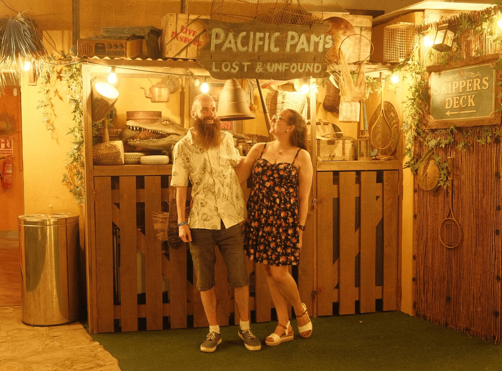 Tom Lally smiling beside a woman in a floral dress in front of a rustic, lantern-lit wooden booth