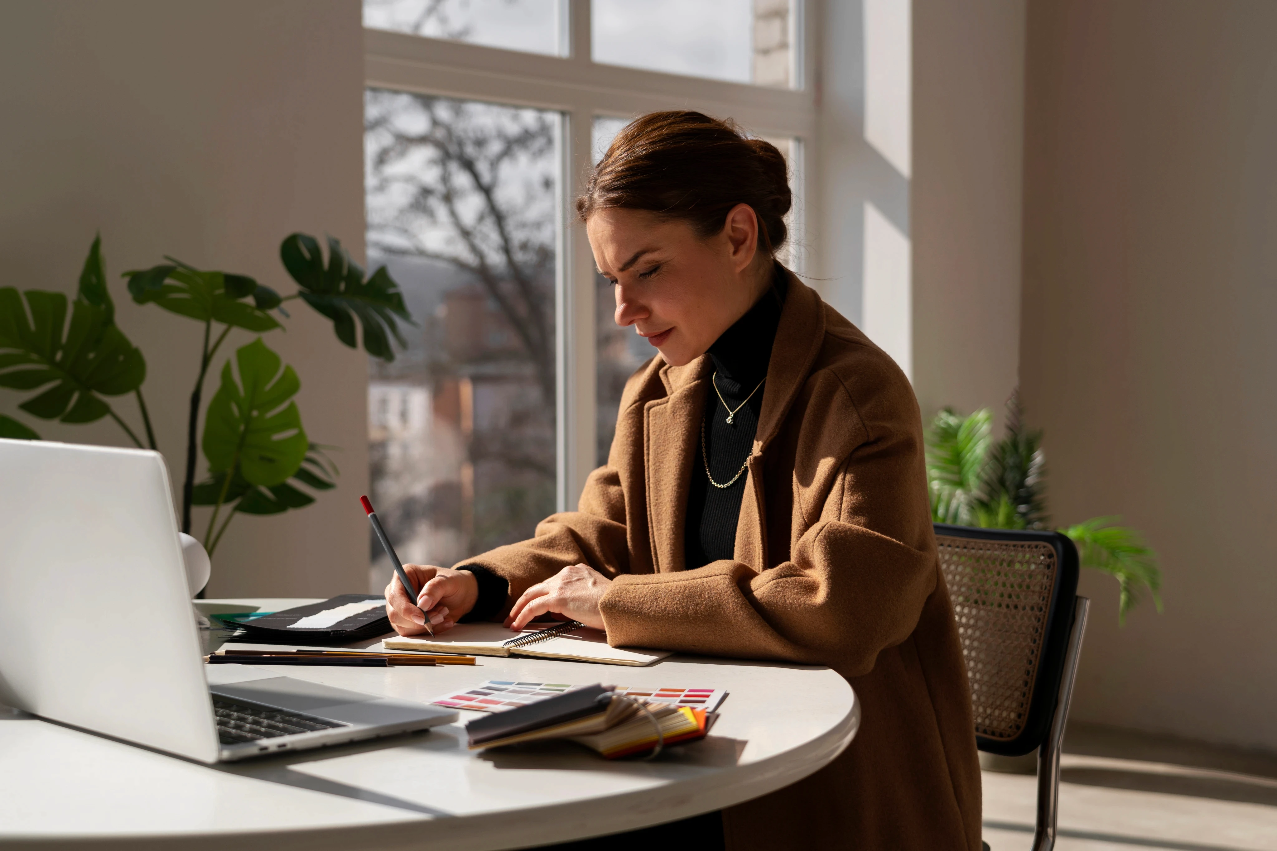 A woman in a brown coat works intently at a desk with a laptop and notepad. Sunlight streams through large windows, and green plants add a calming touch.