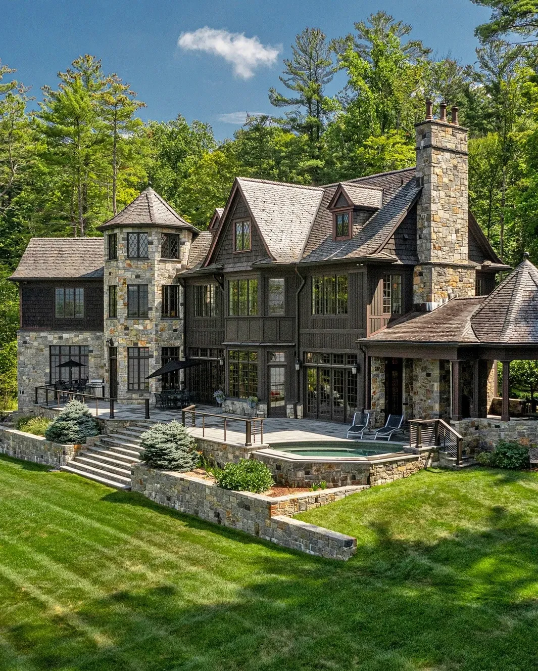  Rustic luxury home with a stone and wood exterior, featuring a steeply pitched roof, multiple chimneys, a turret, large windows, a stone patio with seating, a small hot tub, and lush green lawn surrounded by dense forest under a clear blue sky.