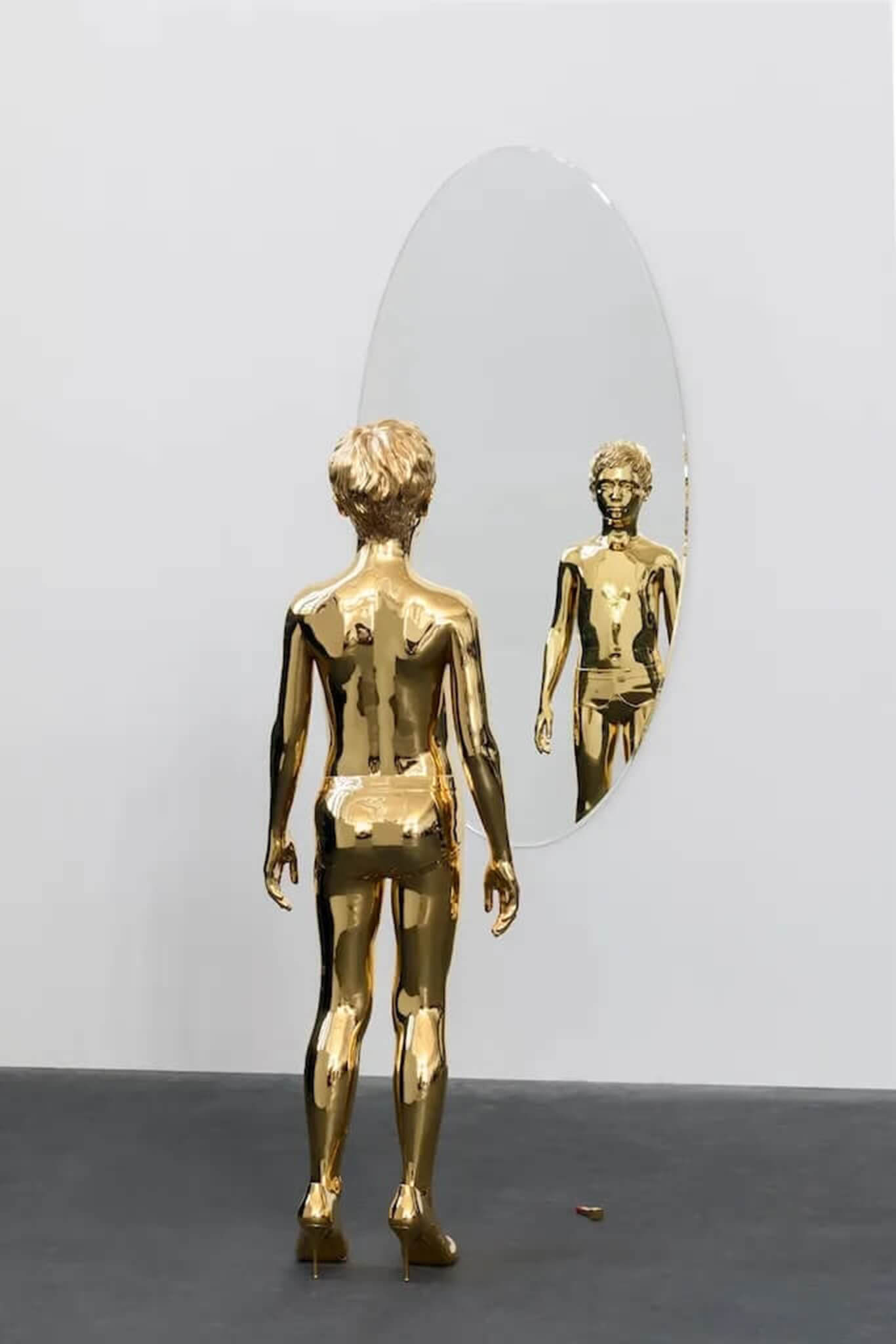 Gold human figure sculpture in high heels facing an oval mirror, reflecting its face against a minimalist white wall.
