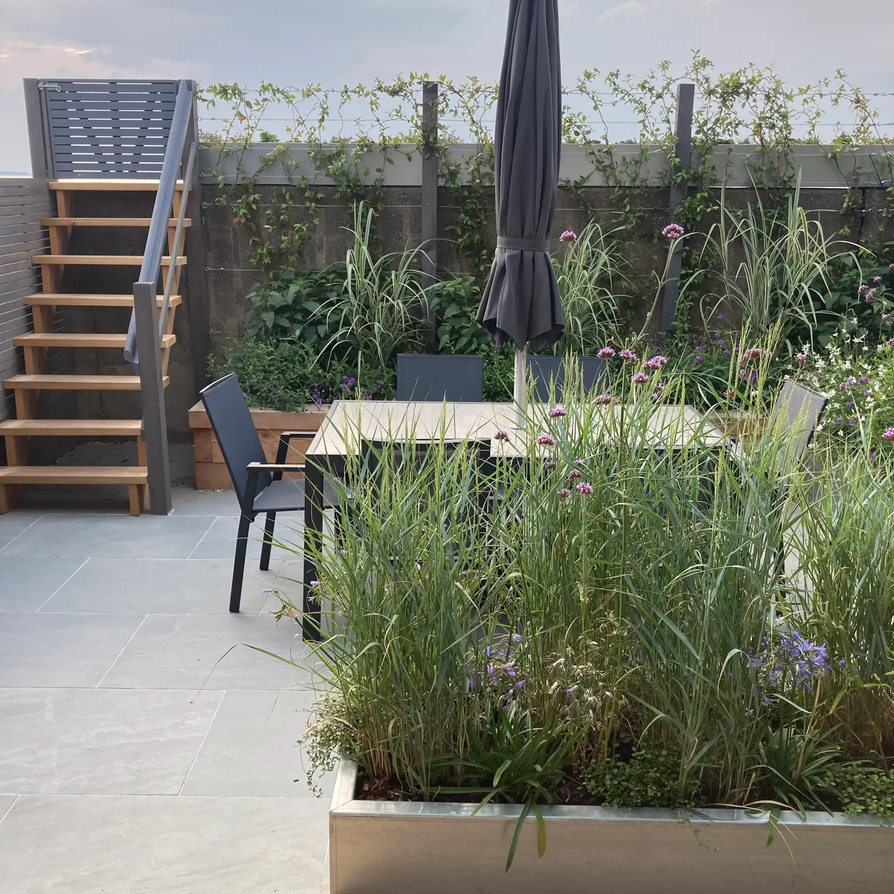 A peaceful garden with a wooden table and chairs, surrounded by greenery and a staircase in the background.