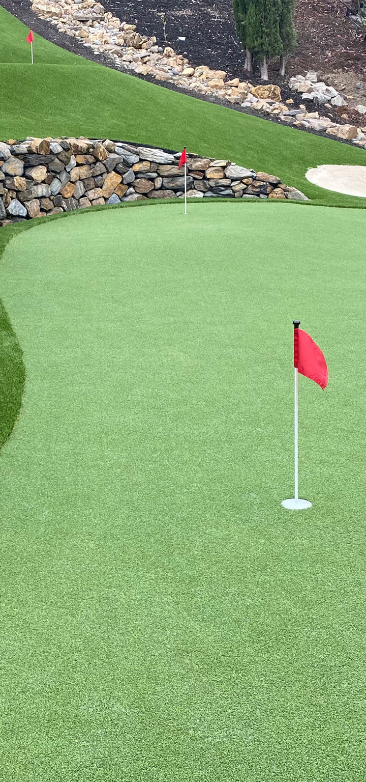 Backyard putting green with red flags and a sand bunker, edged by a curved natural stone wall.
