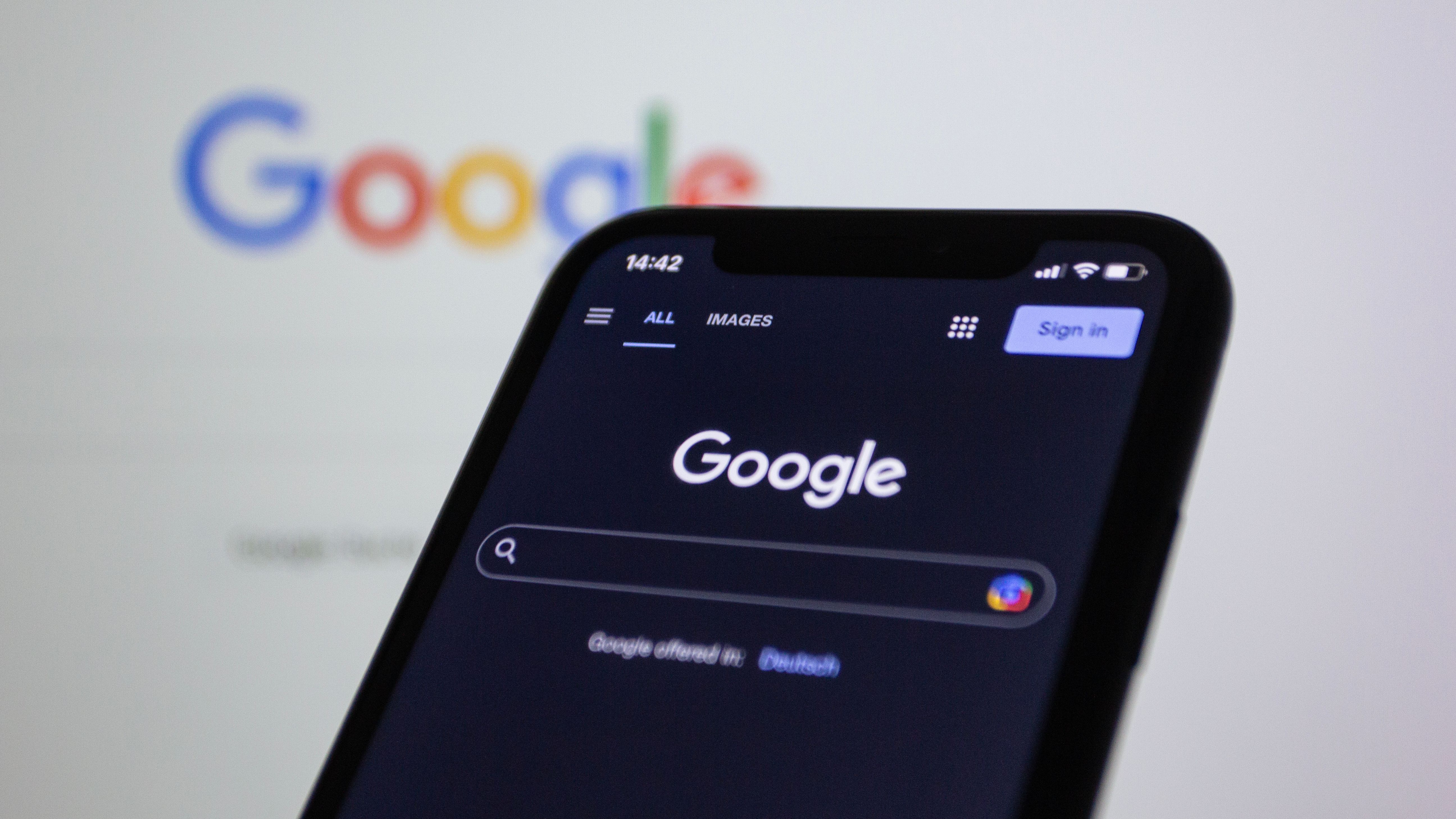 A smartphone displaying Google's search page in dark mode is held in front of a blurred computer screen showing the colorful Google logo, highlighting the search engine's interface and branding.