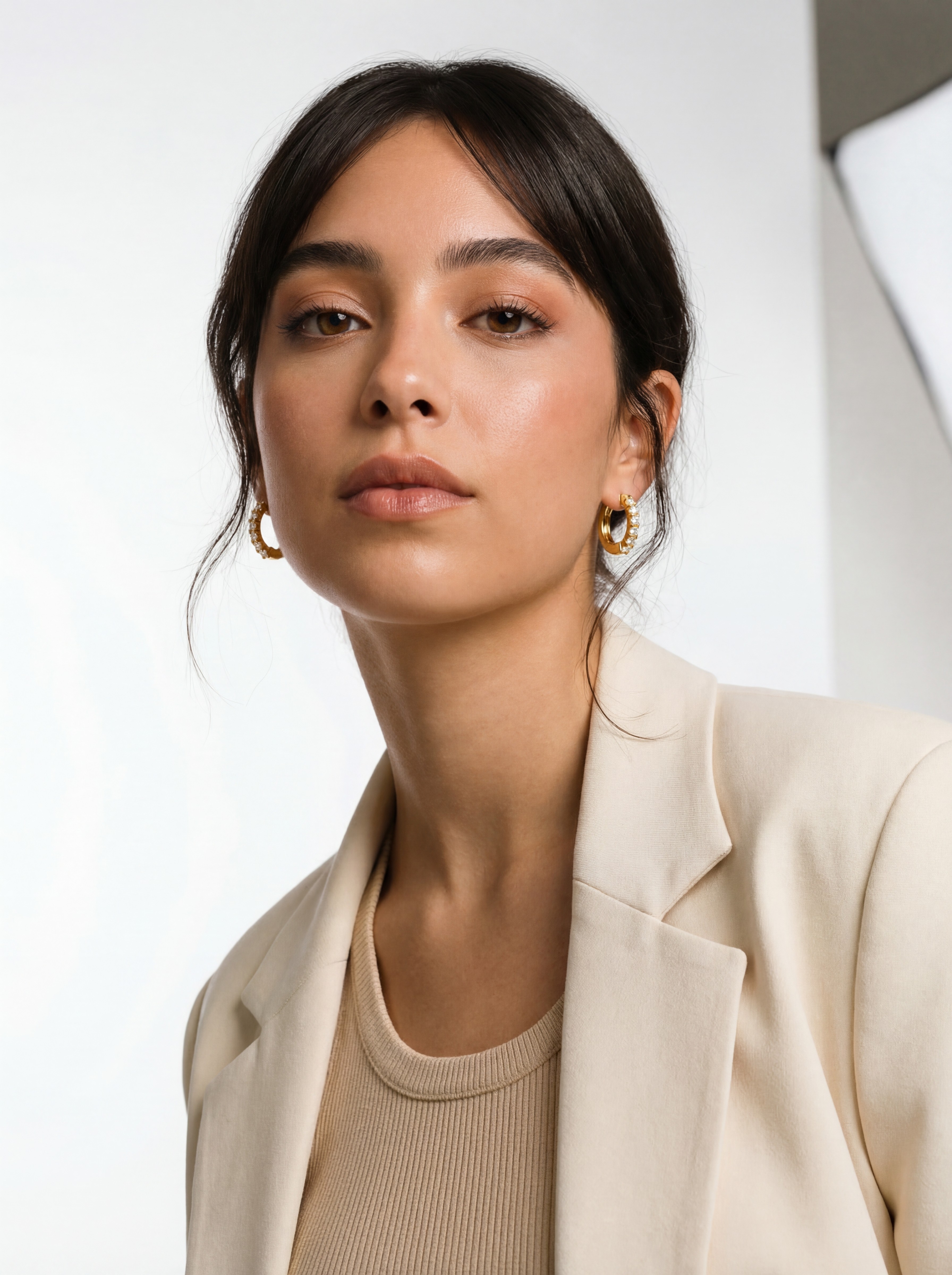 Isabella Vega AI model wearing gold huggie hoop earring, bright editorial studio, cream blazer elegant updo, Gemzy AI jewelry studio